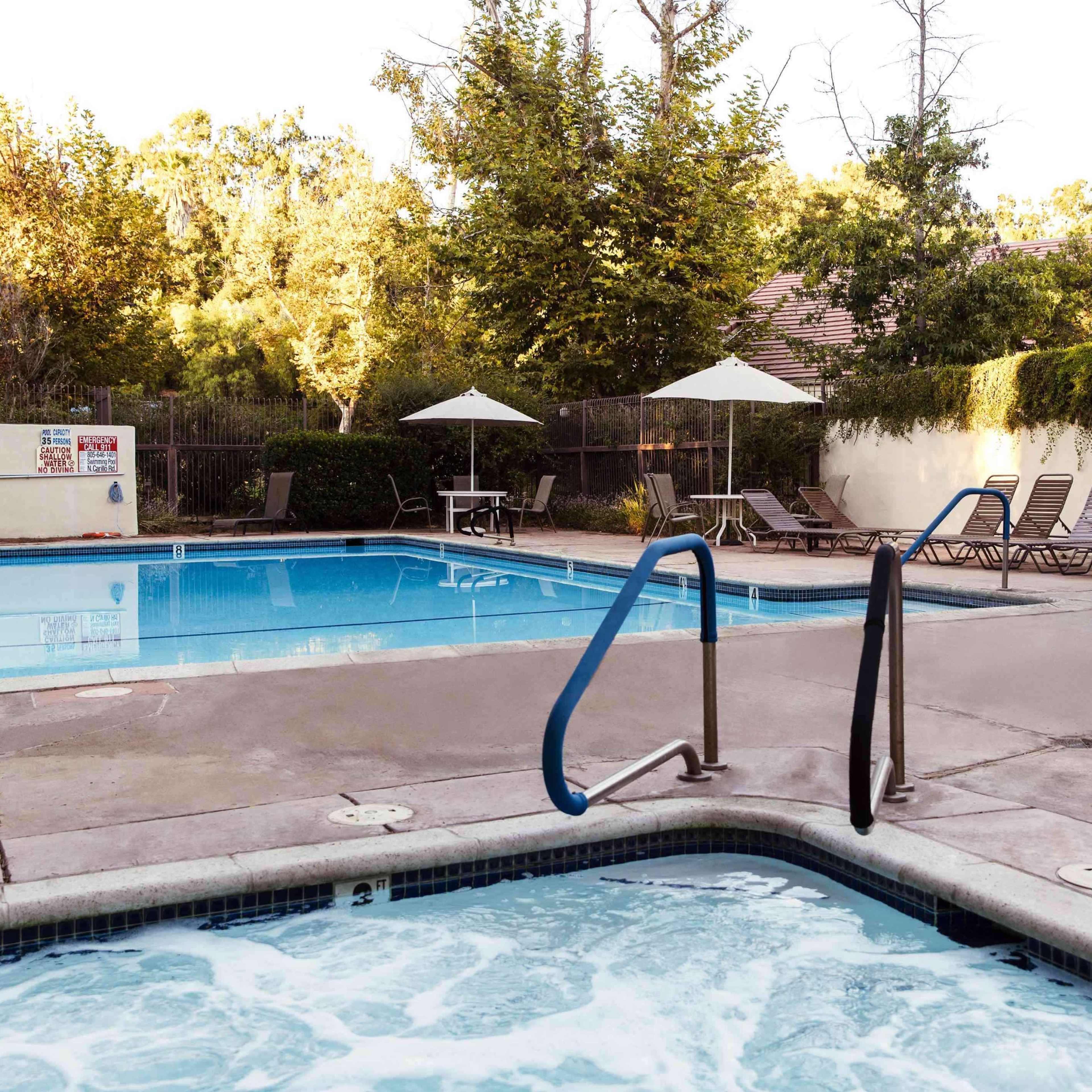 A swimming pool area featuring a main pool, a hot tub, lounge chairs, and shaded umbrellas surrounded by greenery.
