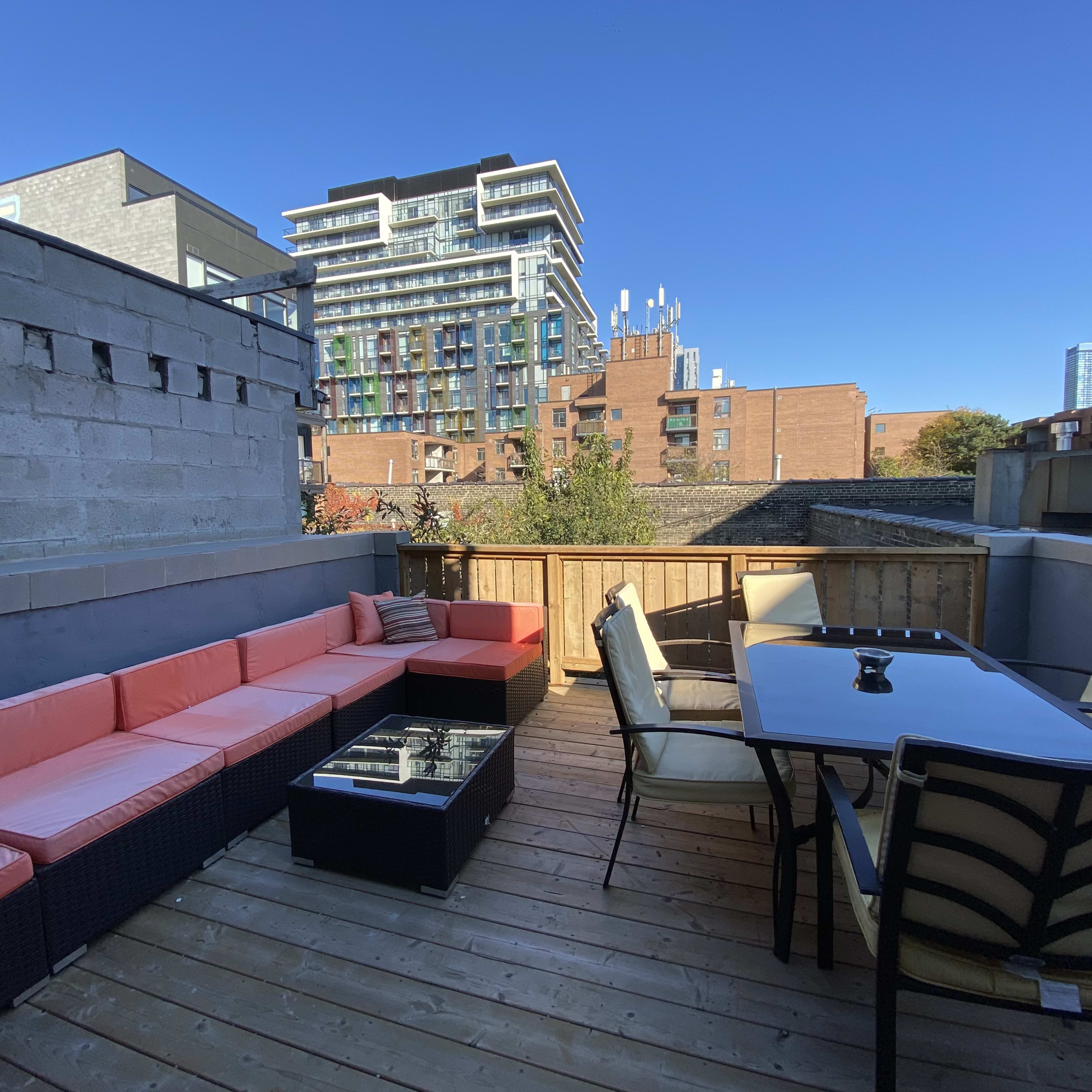 A balcony with a wooden deck featuring a seating area with orange cushions, a glass-topped table, and a view of buildings in the background.