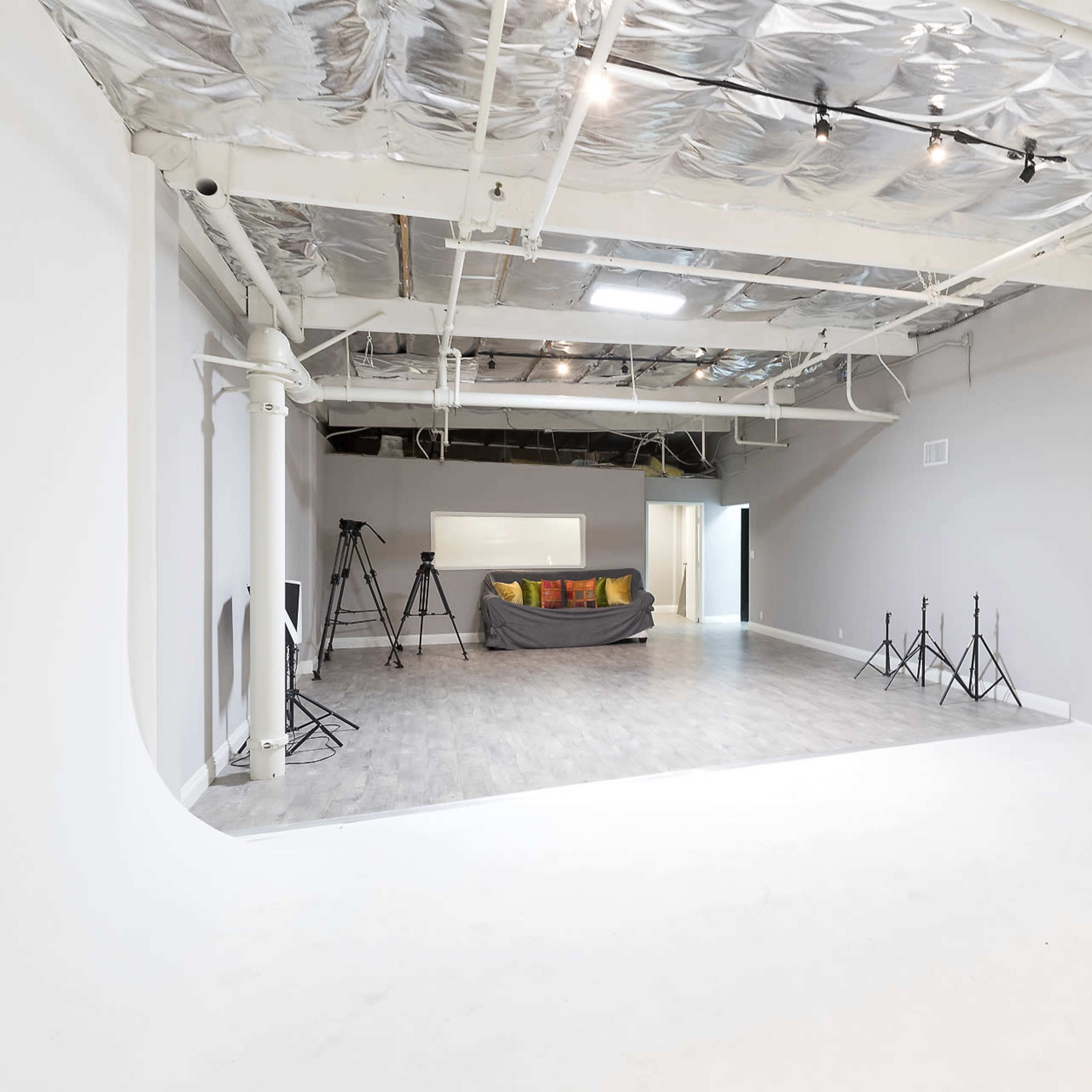 A spacious, unfinished studio with a light-colored floor, exposed ceiling, and several standing tripods in the background.