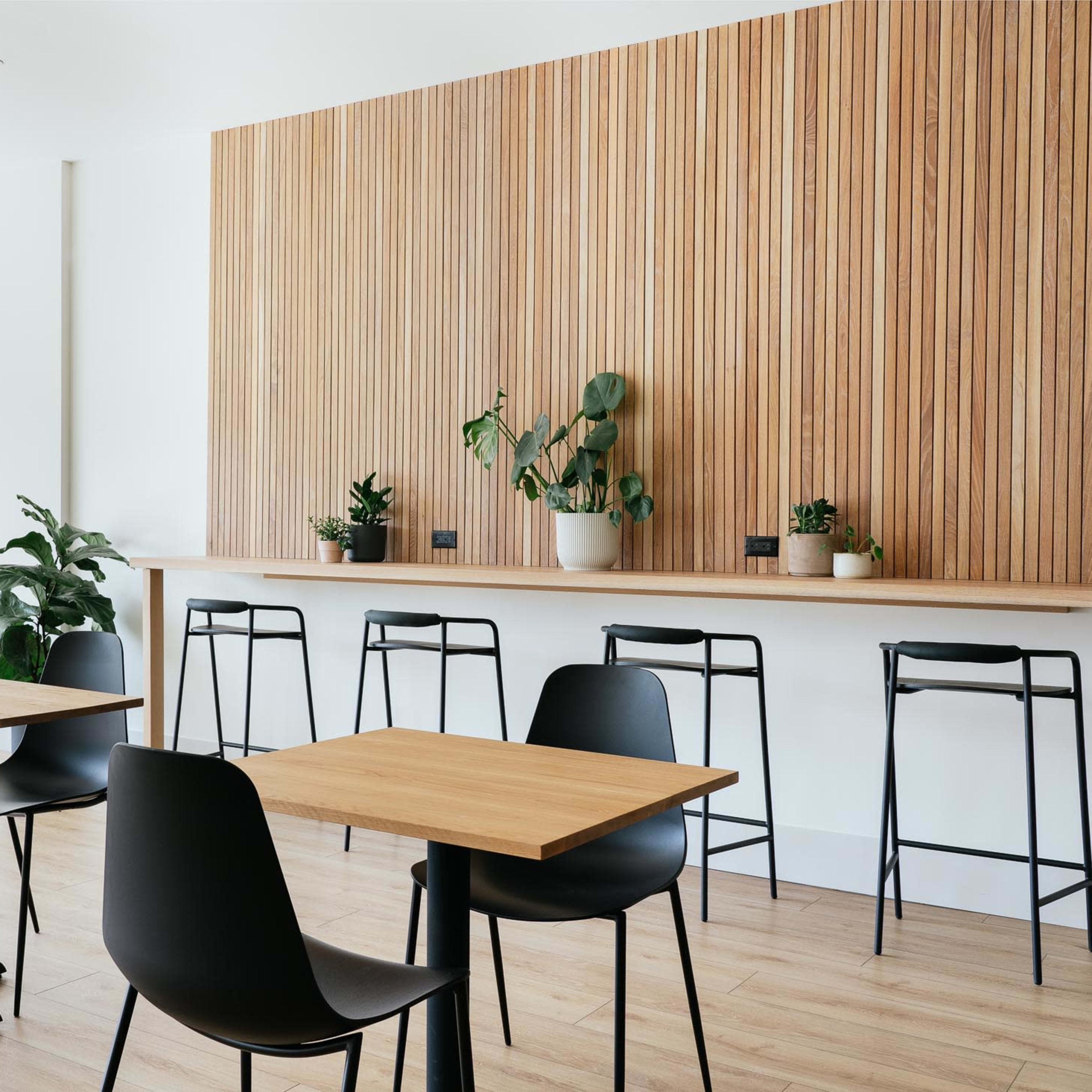 A modern café interior with wooden walls, minimalist furniture, and plants arranged on tables and a counter.