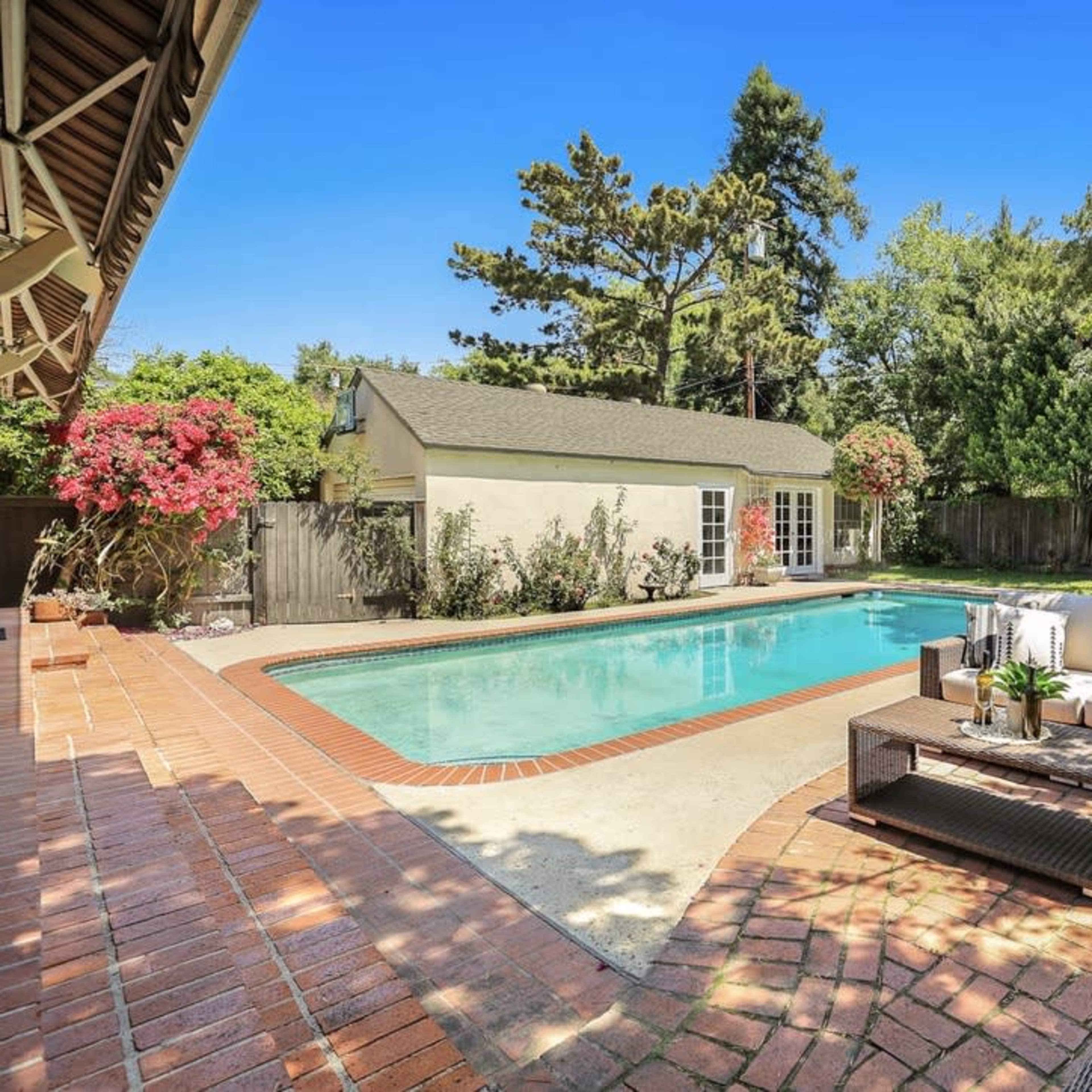A backyard features a rectangular pool surrounded by a brick patio and a seating area with a sofa, overlooking a well-maintained garden.