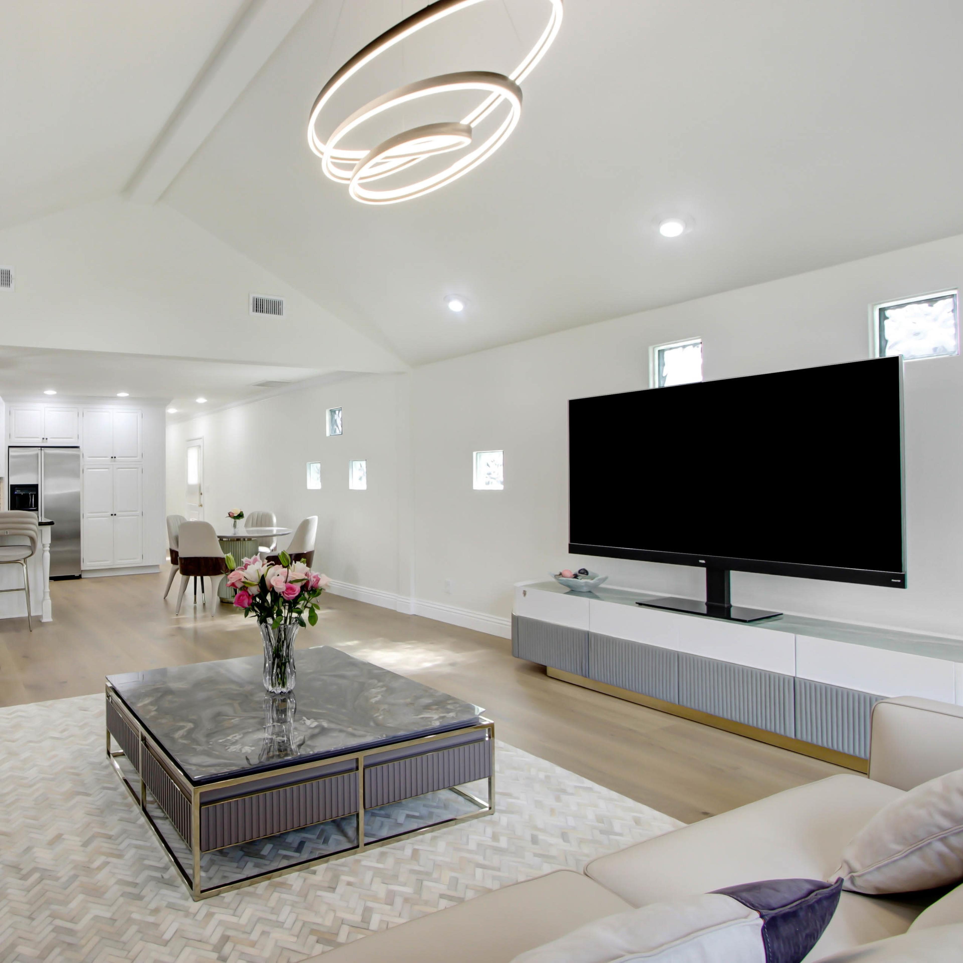 A modern living room with a beige sofa, a glass coffee table, and a large television, set in an open layout adjacent to a kitchen and dining area.