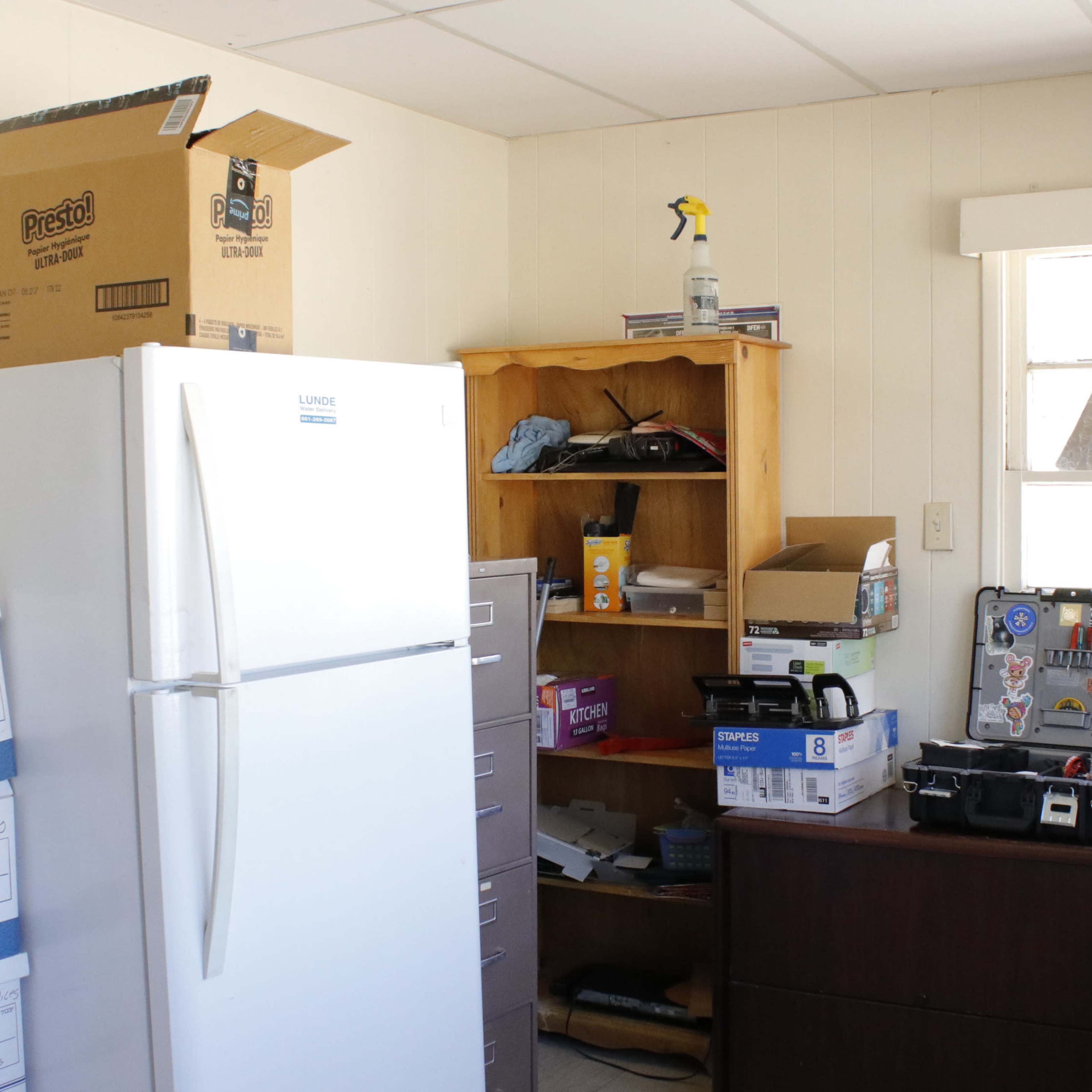 A cluttered room features a white refrigerator, a wooden shelf filled with boxes and containers, and a dark desk with various tools and supplies.