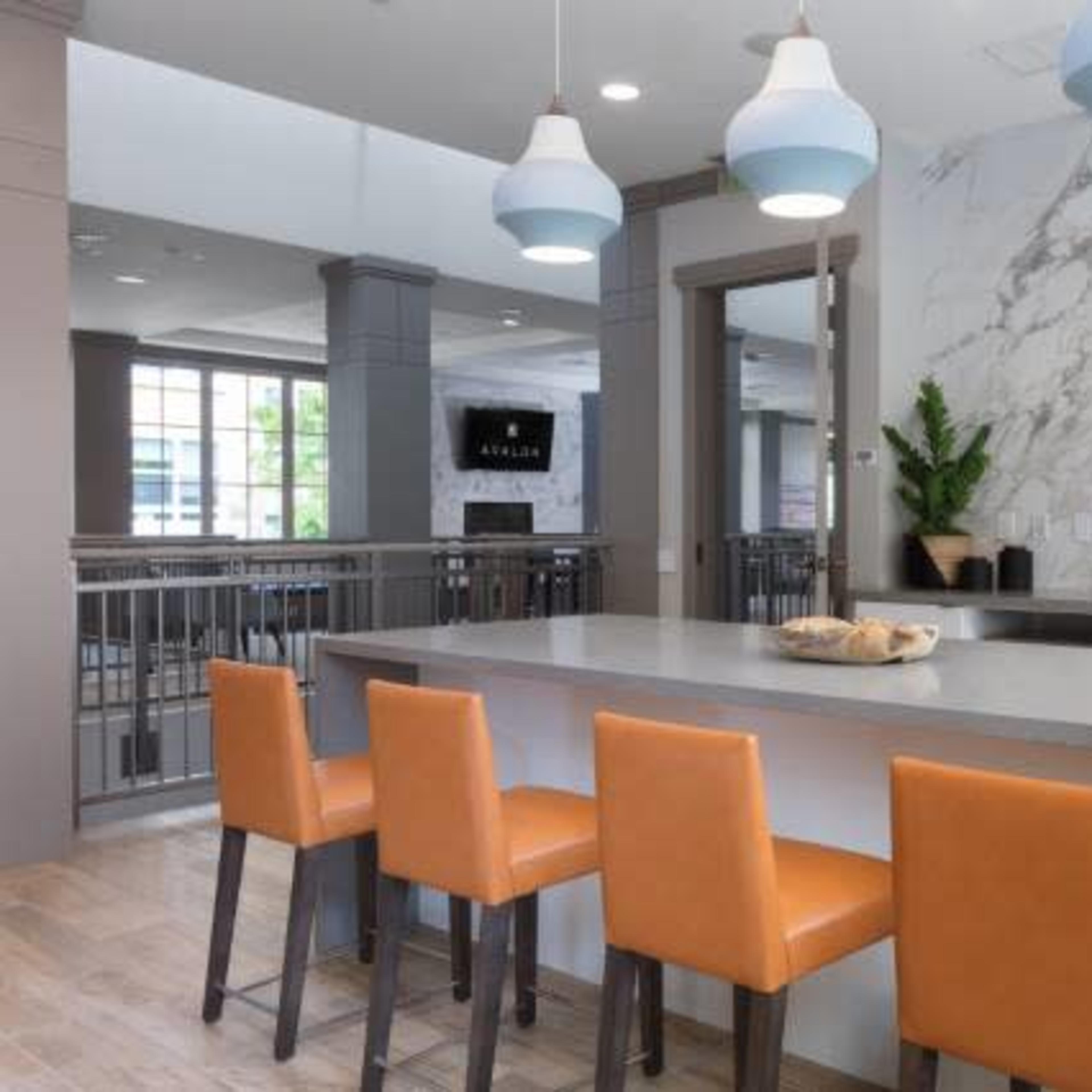 A modern kitchen features a long countertop with orange stools and pendant lighting above, set against a marble accent wall.