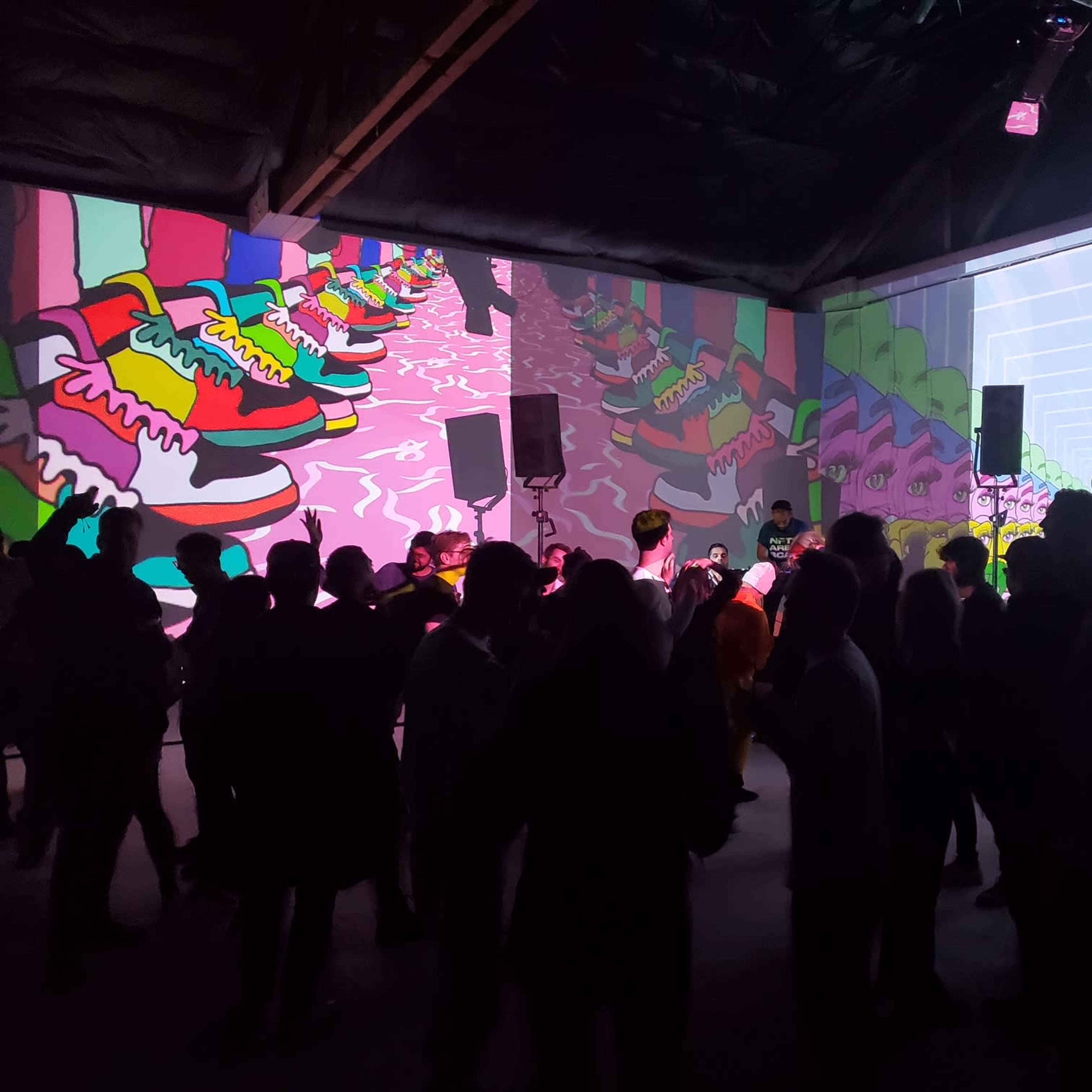 A crowd of people dances in a dimly lit space with colorful projections of sneakers on the walls.