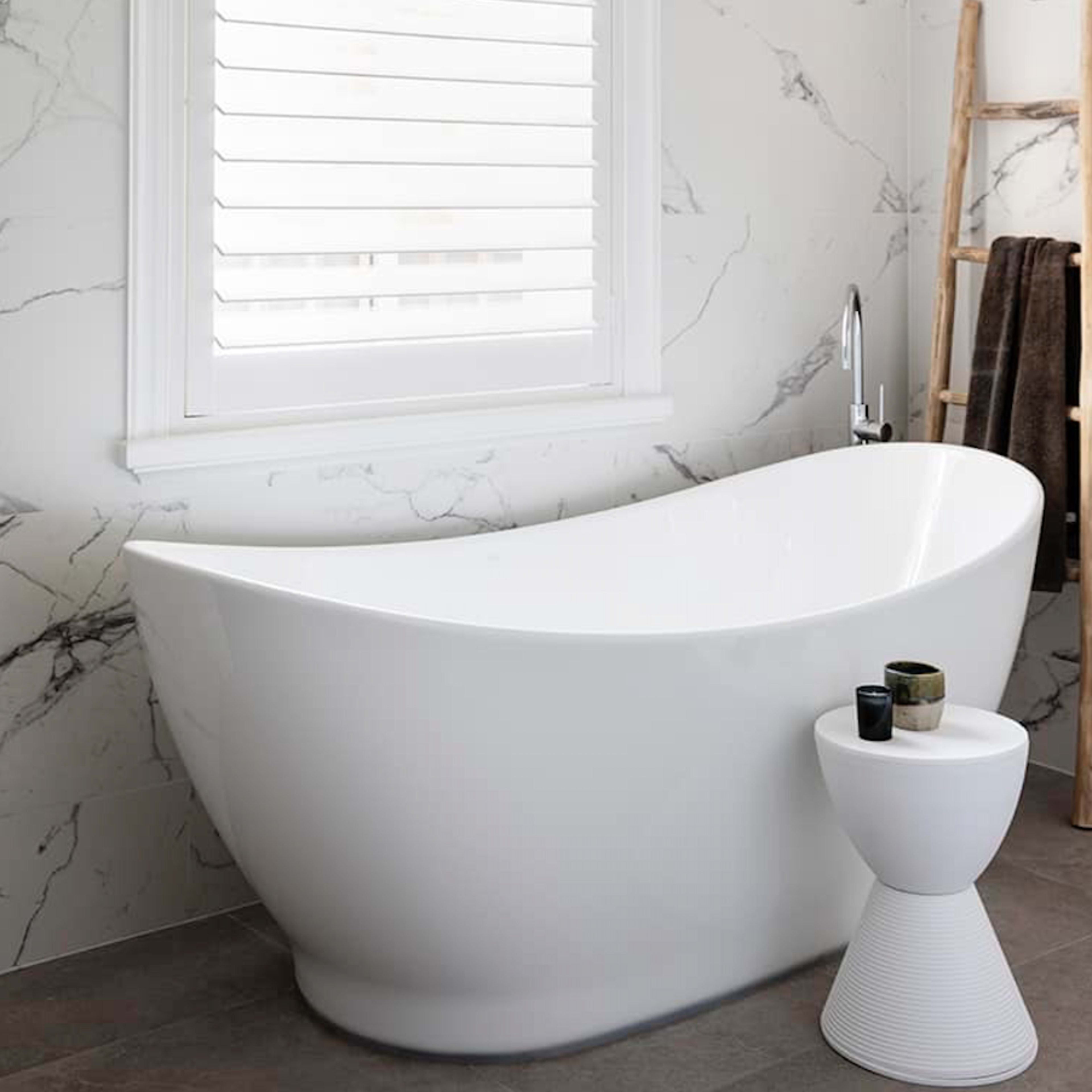 A modern bathroom features a white freestanding bathtub beside a small table, with marble-patterned walls and a window covered by white blinds.