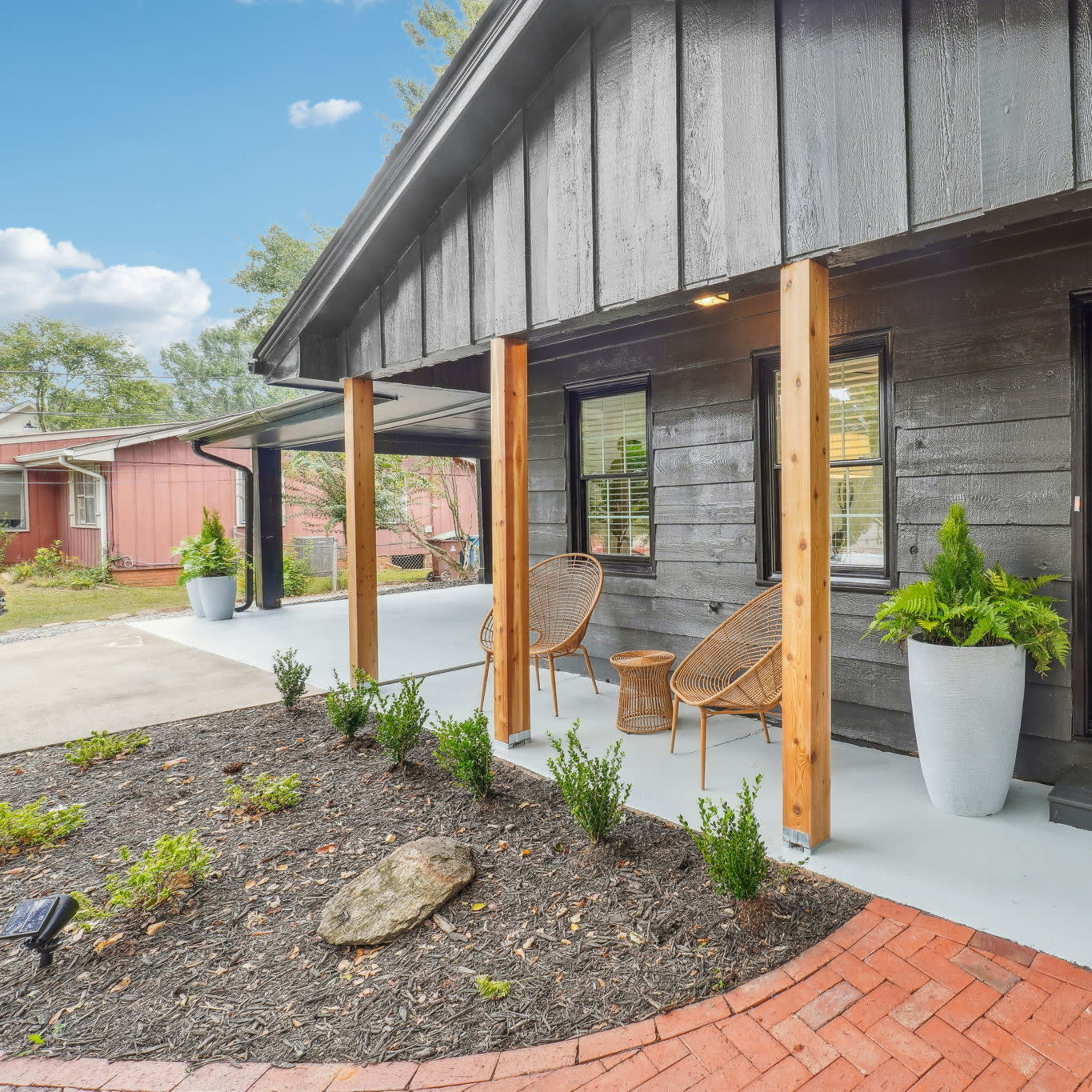 A front porch of a house featuring two chairs, a welcome mat, and large planters with greenery, surrounded by a brick walkway and mulch.