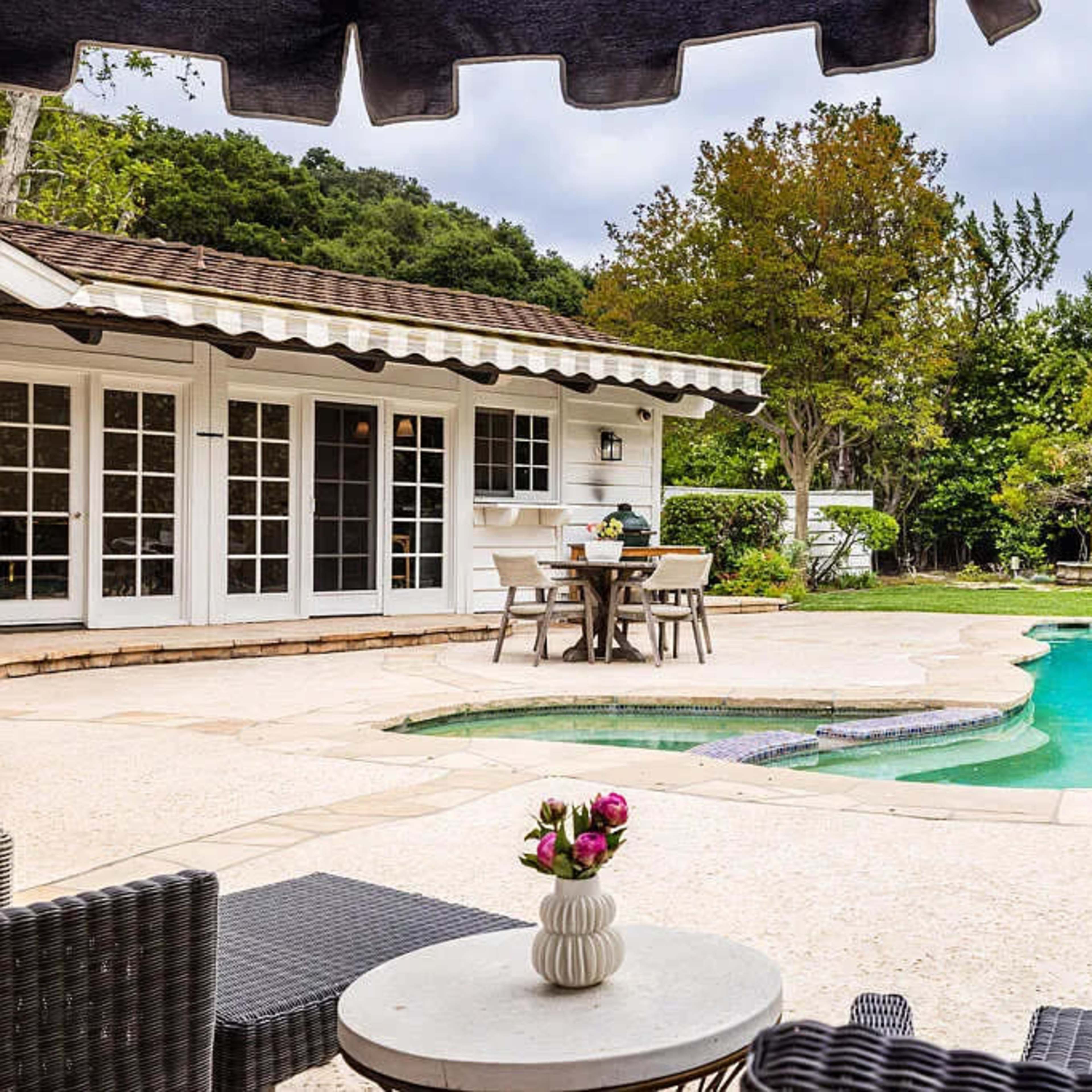 A backyard features a swimming pool surrounded by a patio, dining table, and lush greenery.