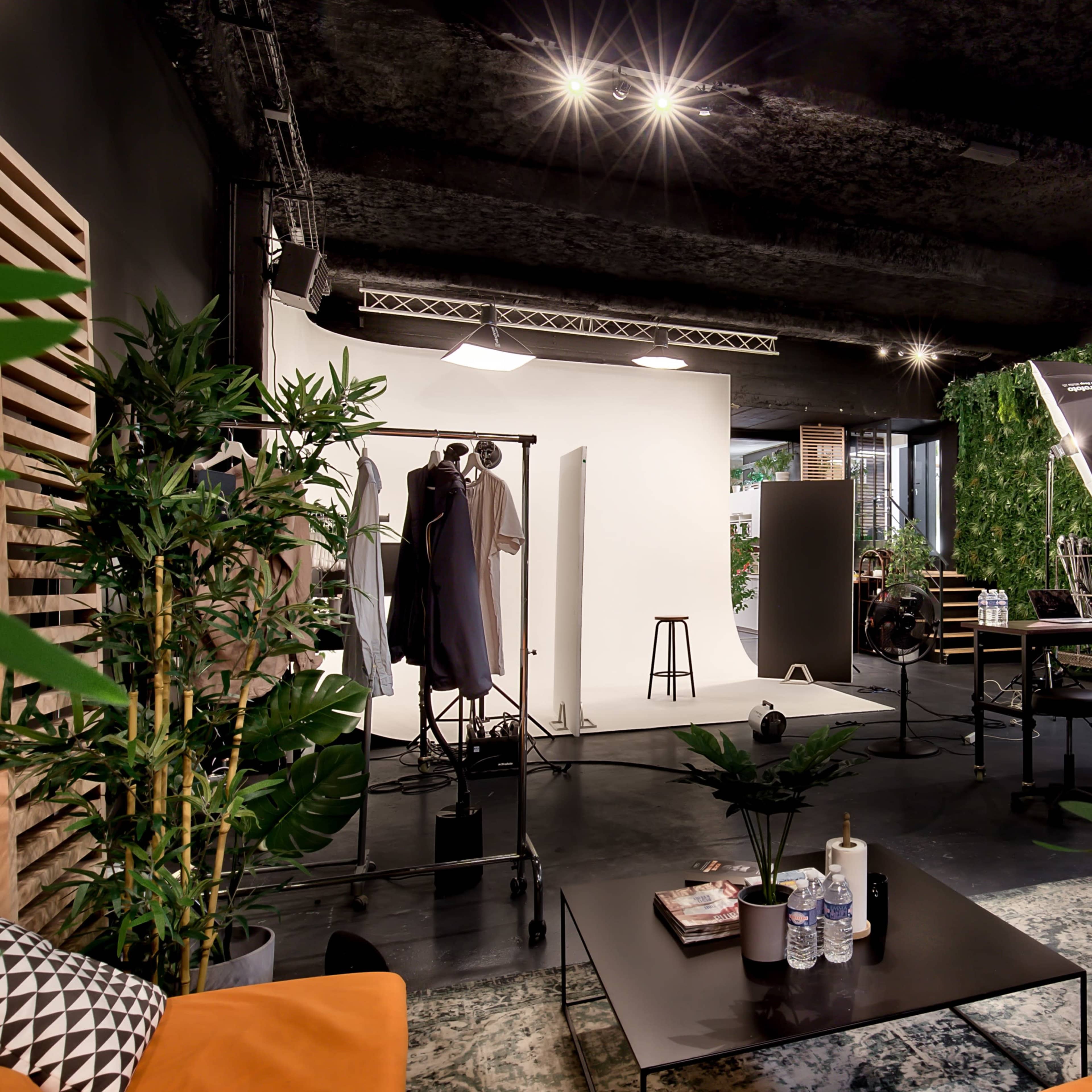 A photography studio featuring a white backdrop, various lighting equipment, and green plants alongside furniture like a table and chairs.