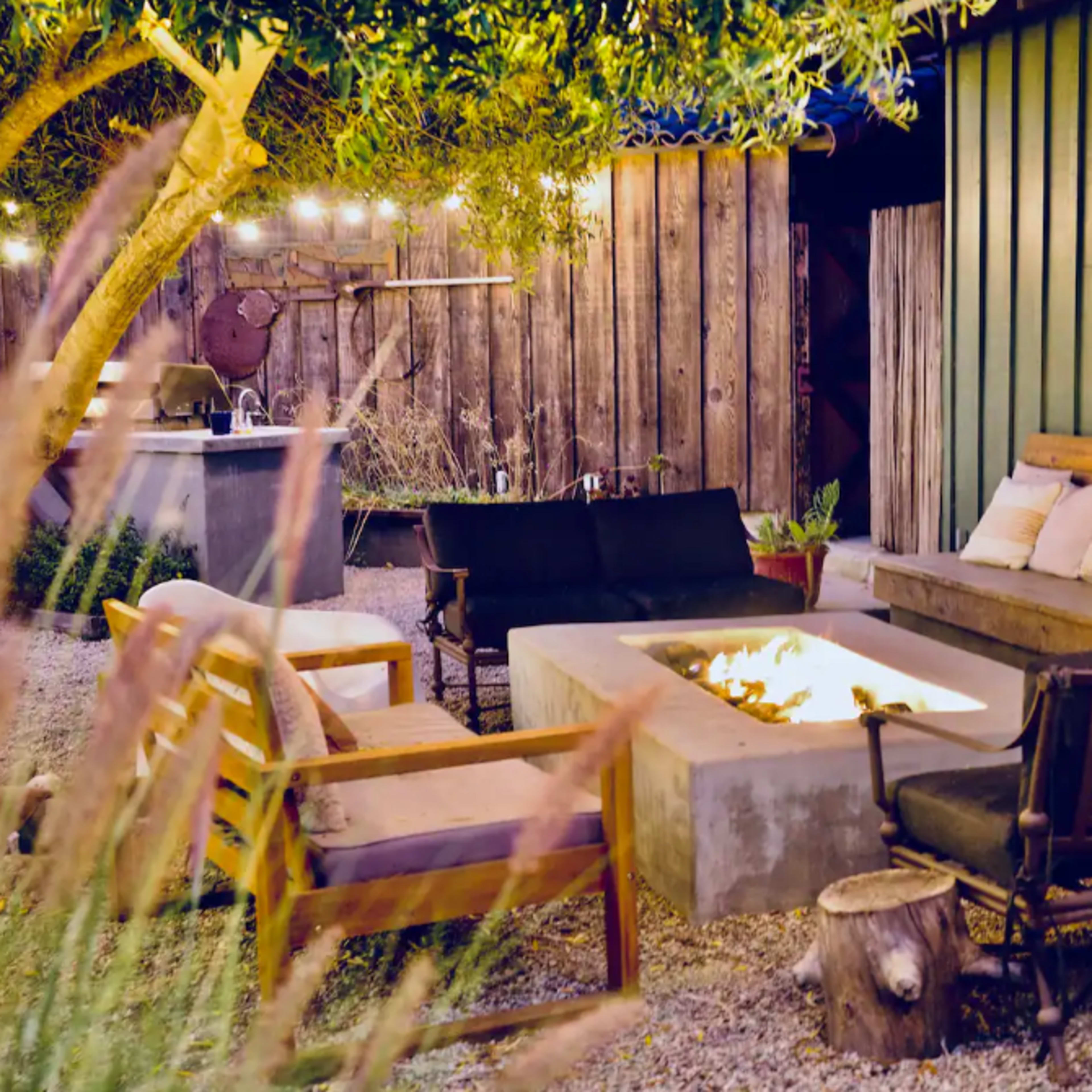 A cozy outdoor seating area featuring a fire pit surrounded by various chairs and a wooden sofa against a green wooden wall.