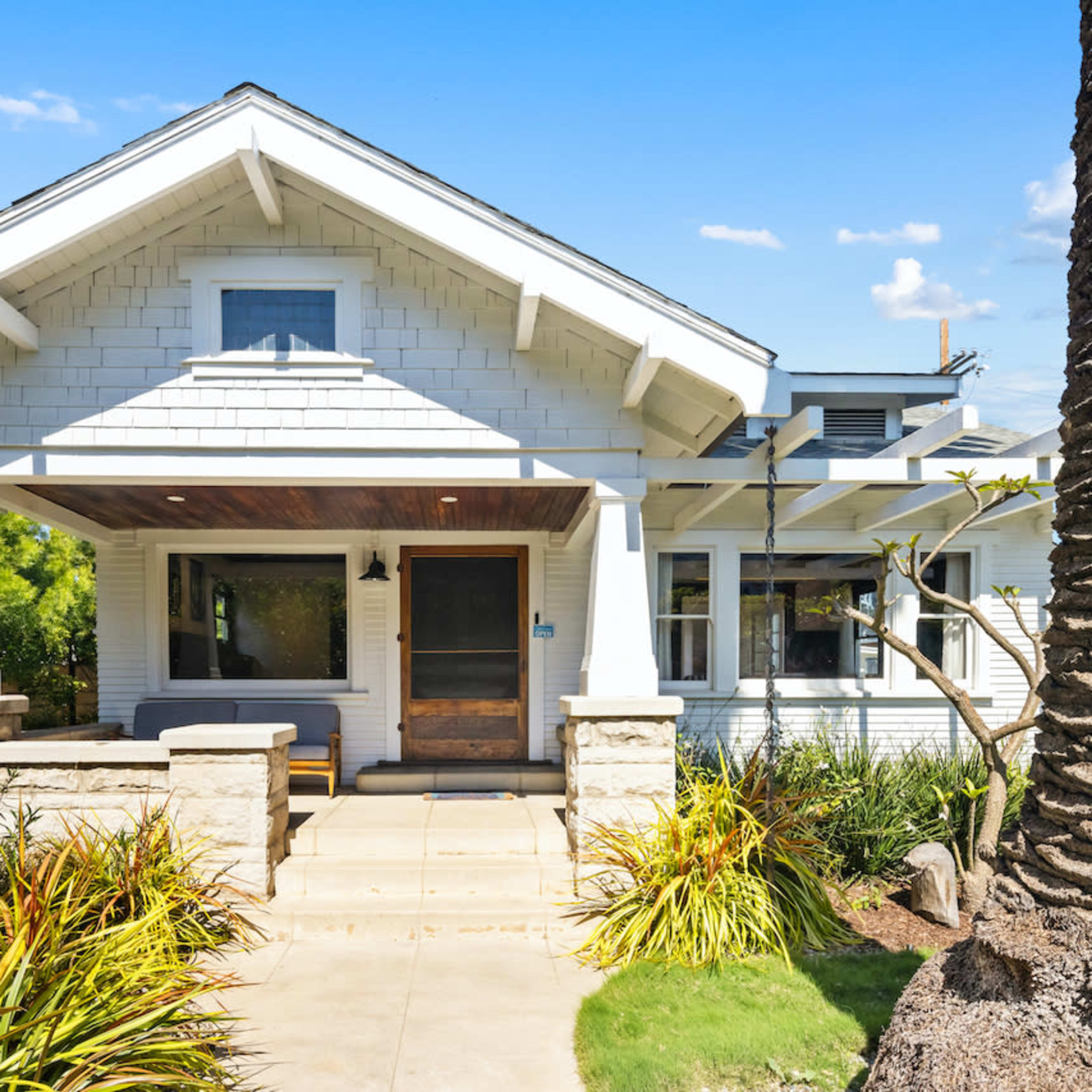 A white craftsman-style house with a front porch, surrounded by landscaped greenery and a palm tree.
