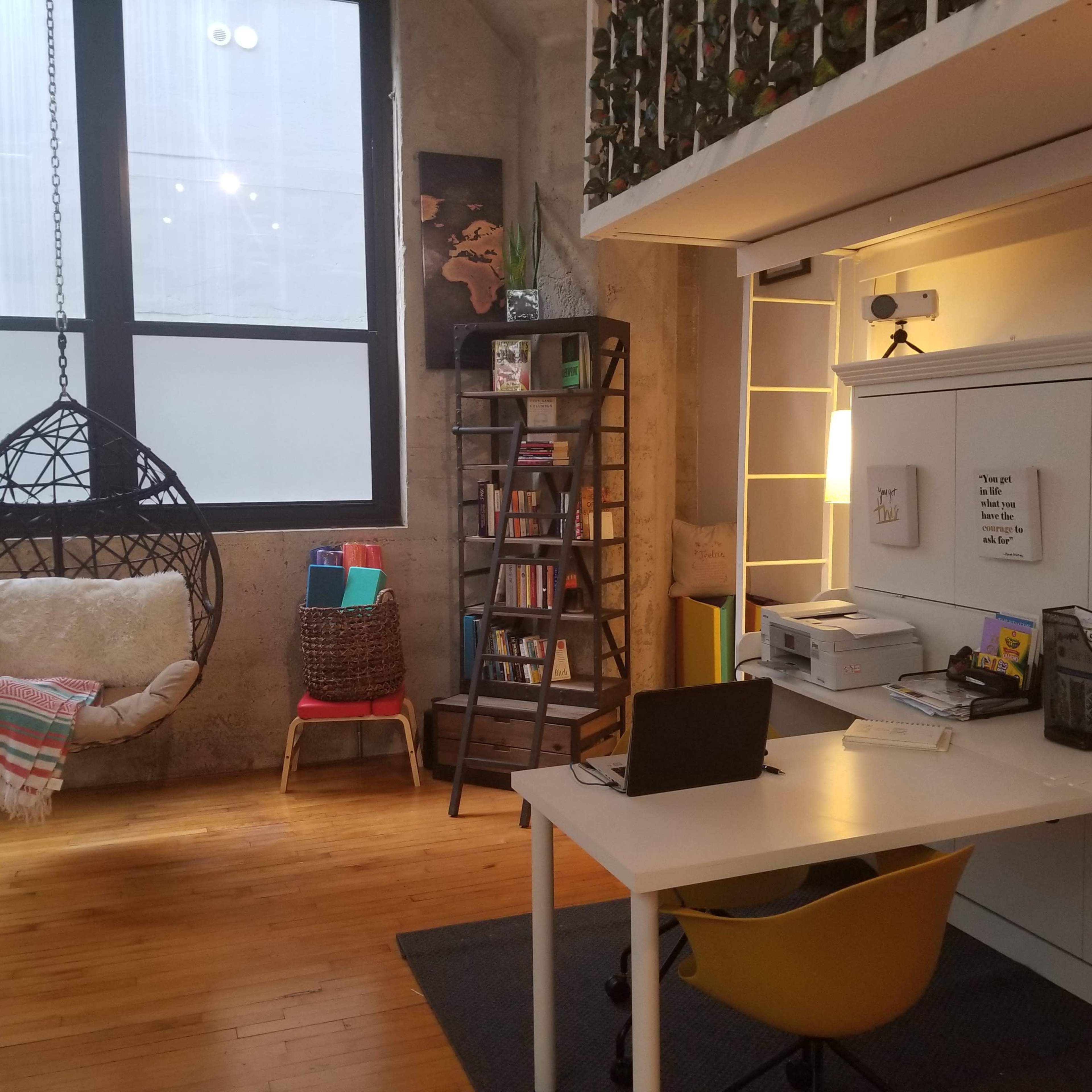 A modern workspace featuring a desk with a computer, a hanging chair, a bookshelf filled with books, and a printer in a well-lit room with large windows.