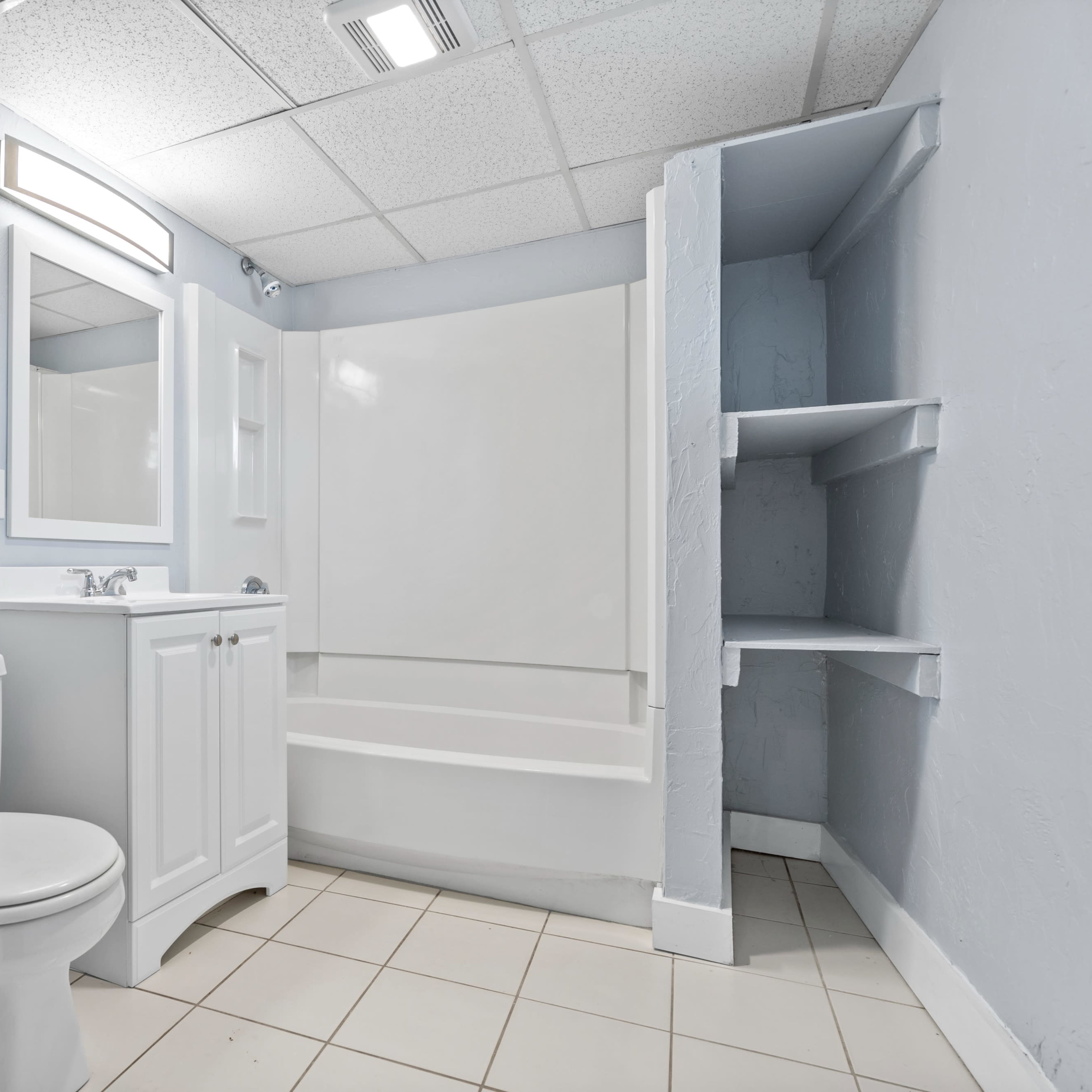 A bathroom features a toilet, a sink with a mirror, a bathtub, and wall-mounted shelves in a light blue environment.