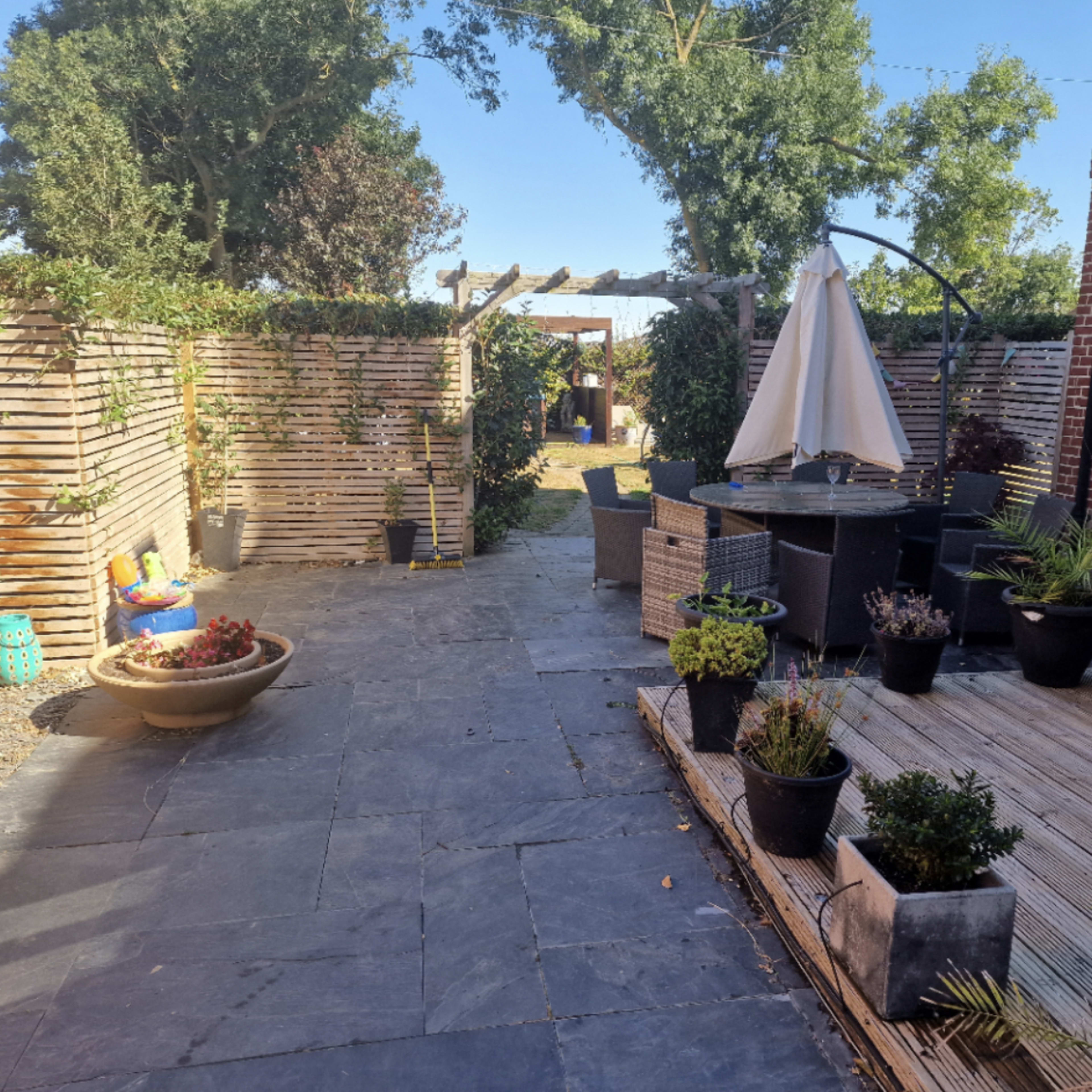 A garden patio area with a circular table and chairs, surrounded by various potted plants, a wooden deck, and a pathway leading to a trellis.