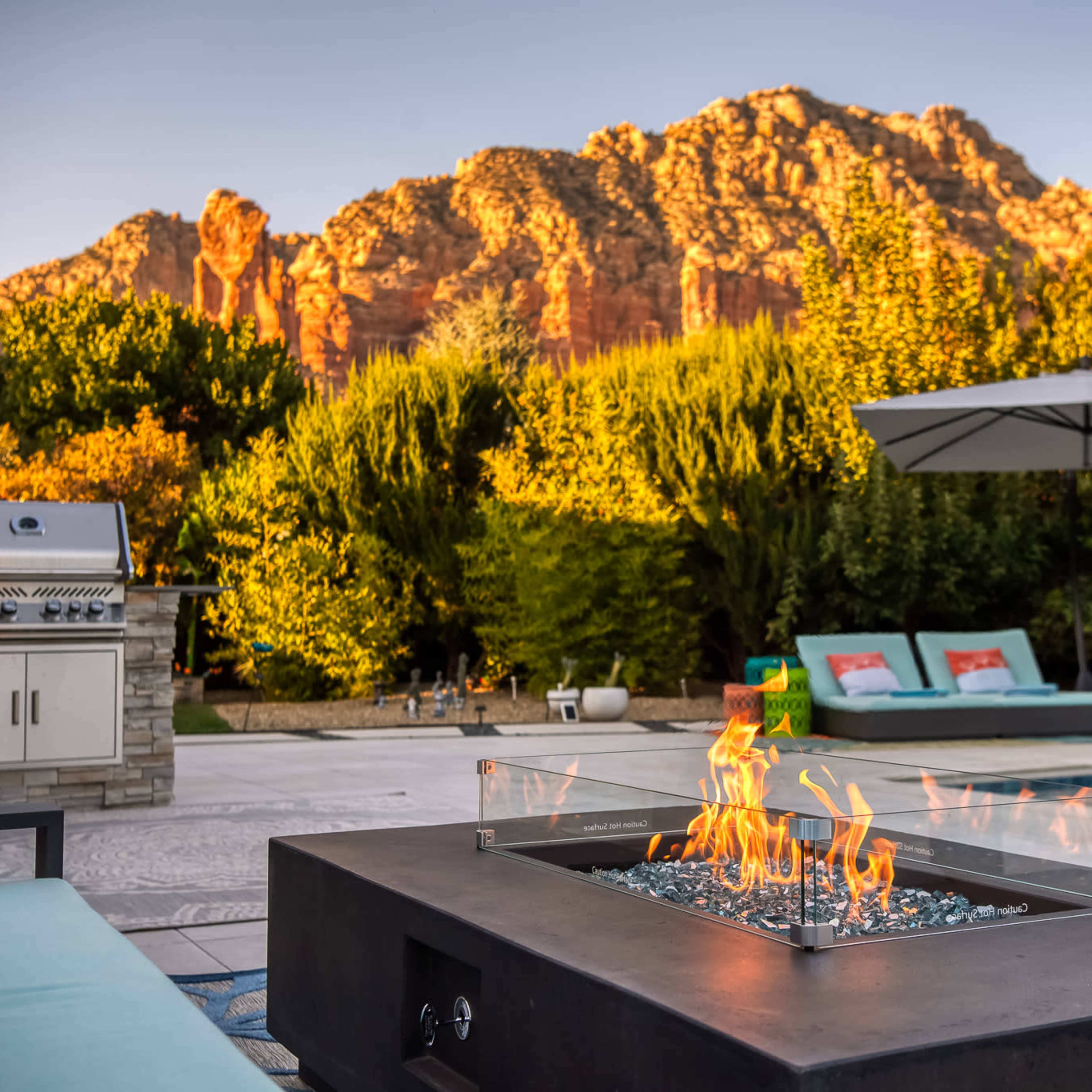 A fire pit with flames is situated in the foreground, surrounded by a stone patio, while a grill and lounge area are visible, with a backdrop of mountainous terrain and greenery.
