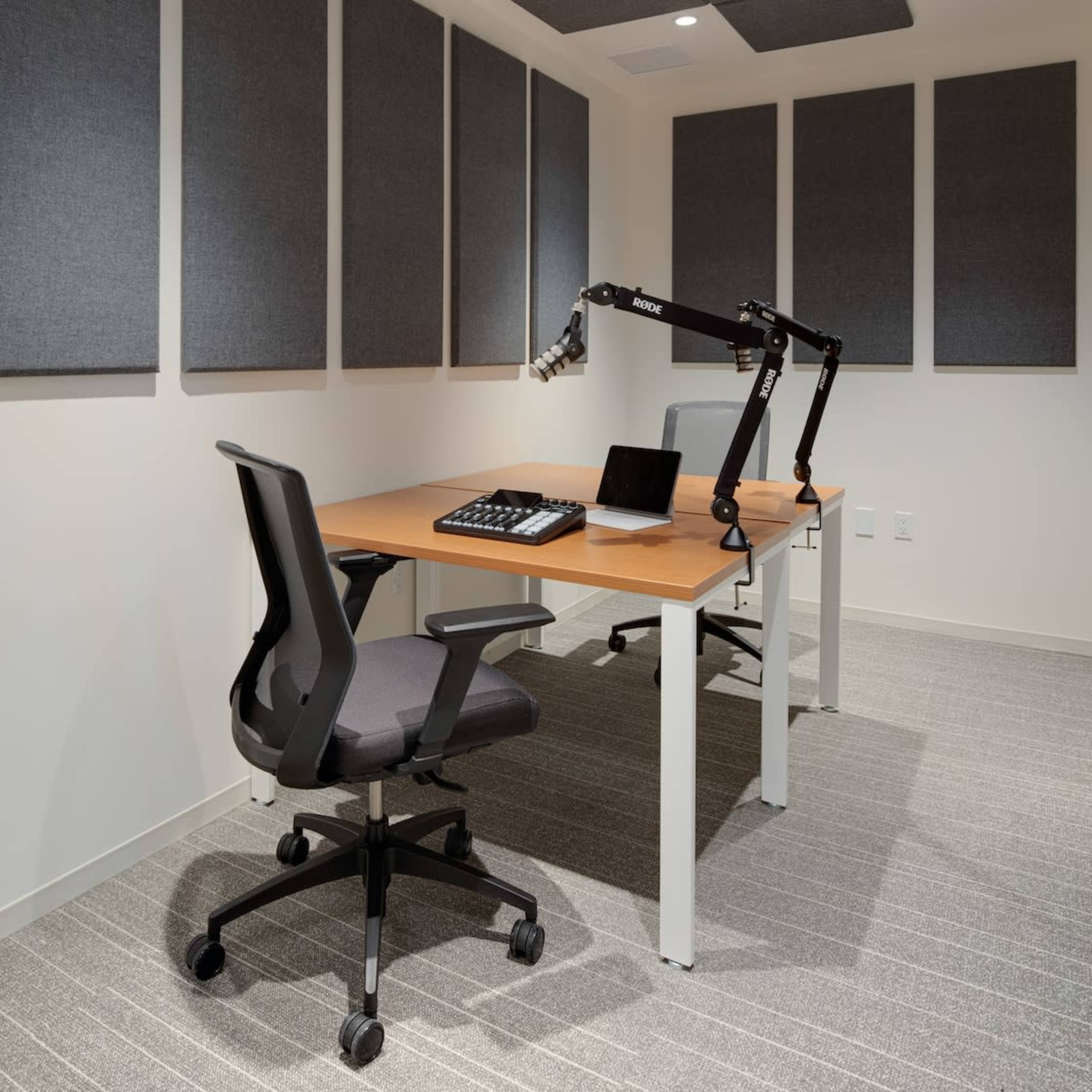 A minimalist office space featuring a large wooden desk, an adjustable microphone arm, a laptop, and acoustic panels on the walls.