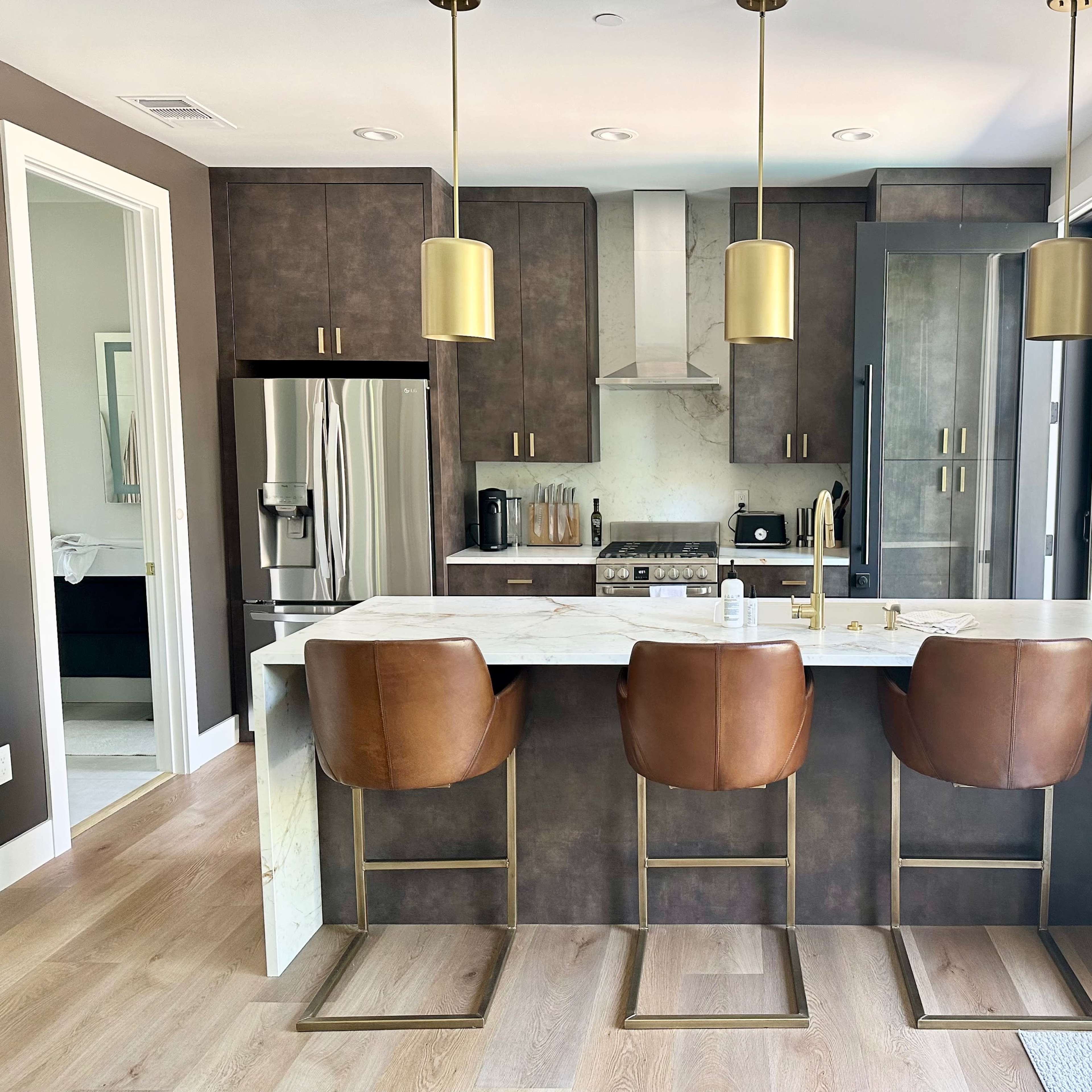 A modern kitchen featuring dark cabinetry, stainless steel appliances, and a marble-topped island with three brown leather bar stools.