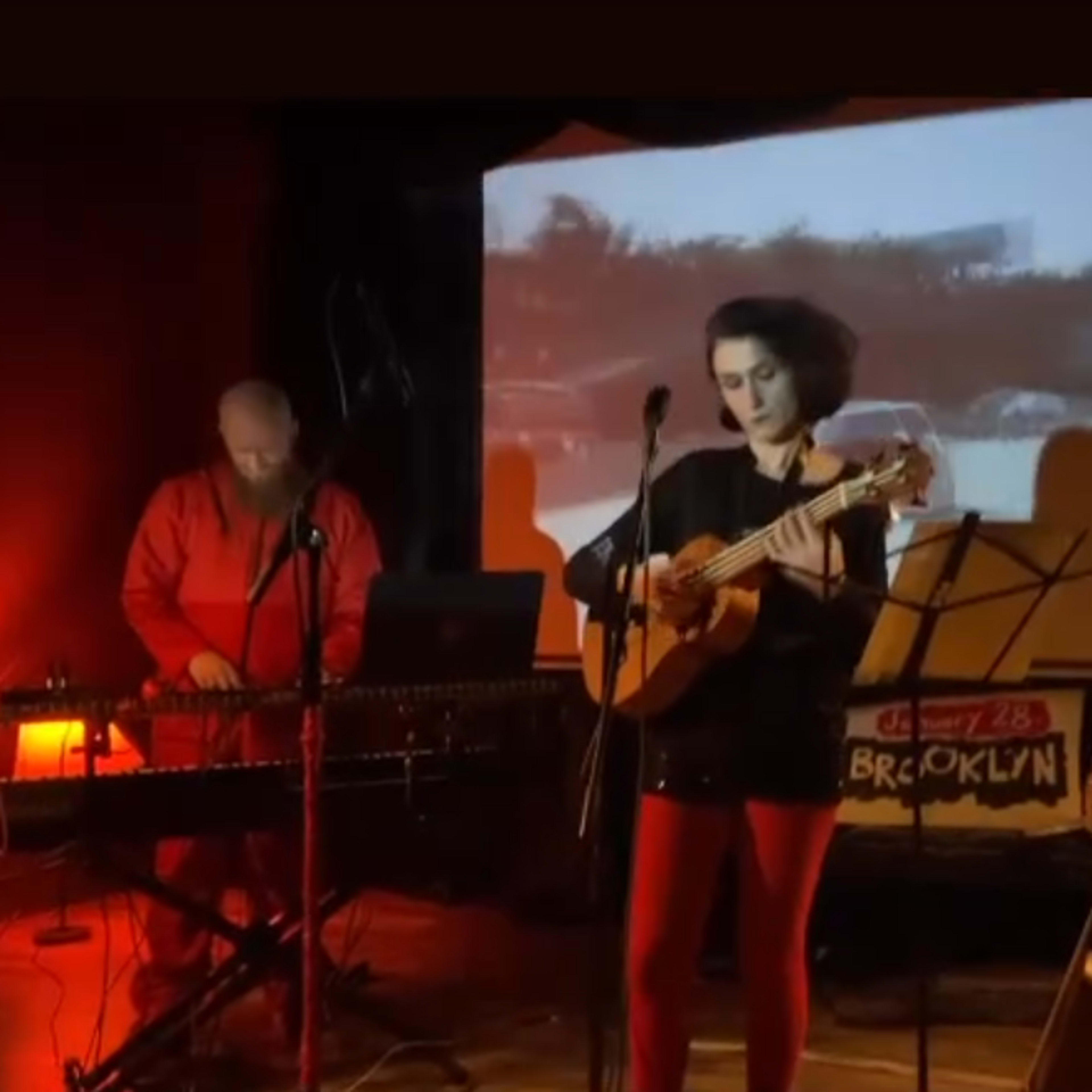 A musician plays a ukulele on stage while a projector displays vintage footage in the background, accompanied by a person on a keyboard.