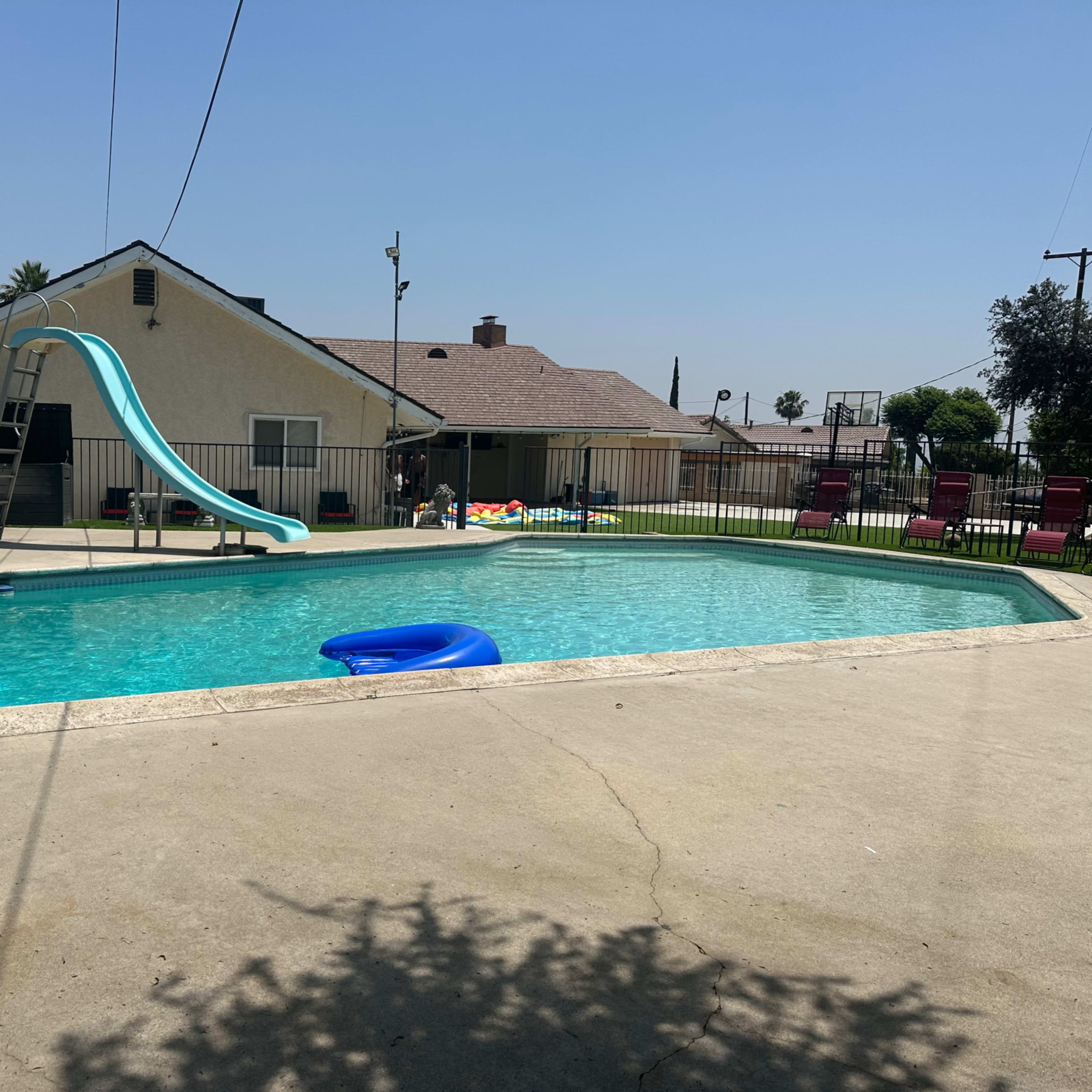 A backyard swimming pool with a slide, surrounded by a fence and various pool floats, in front of a house on a clear day.