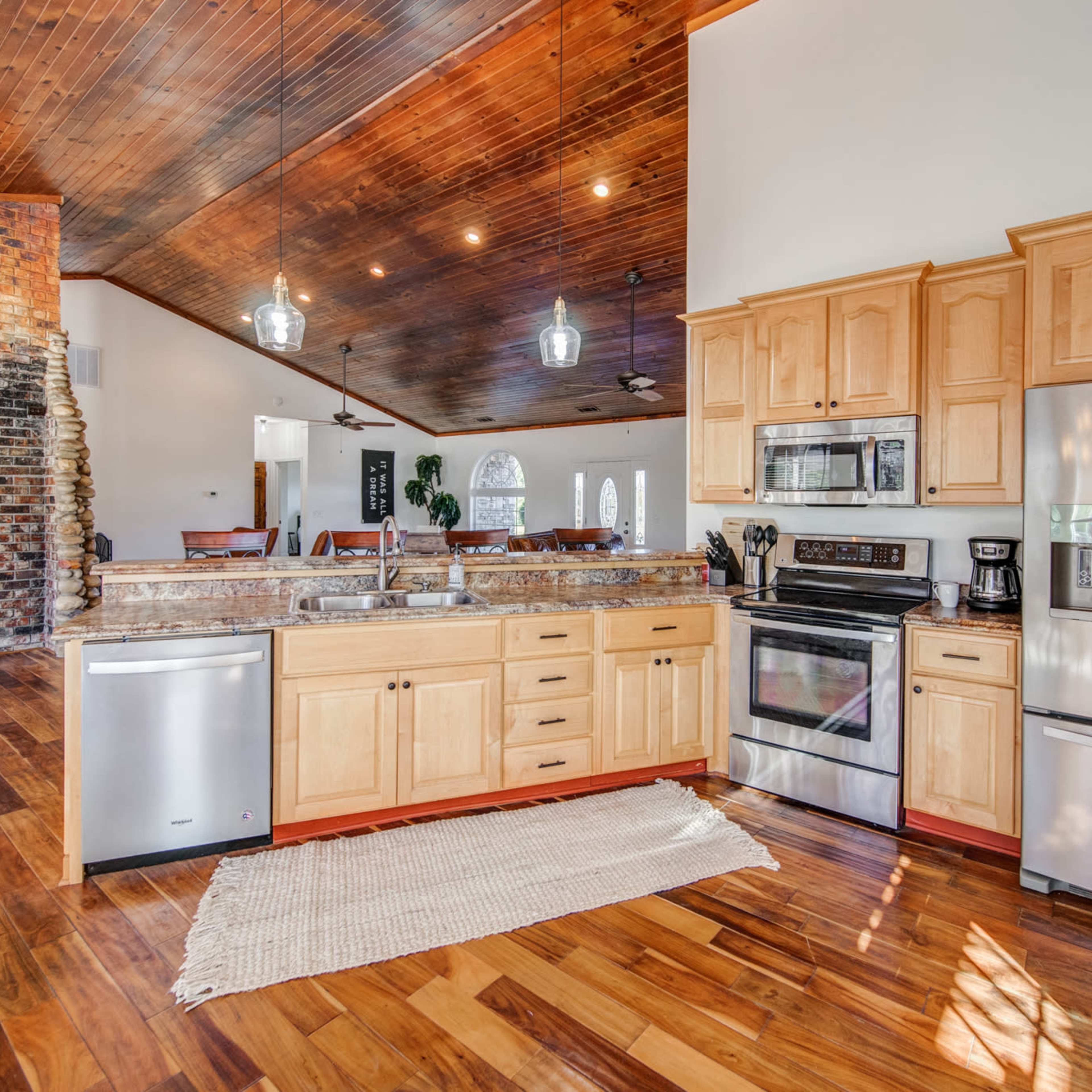 A modern kitchen with wooden cabinets, stainless steel appliances, and a large island with a granite countertop, adjacent to an open living area.