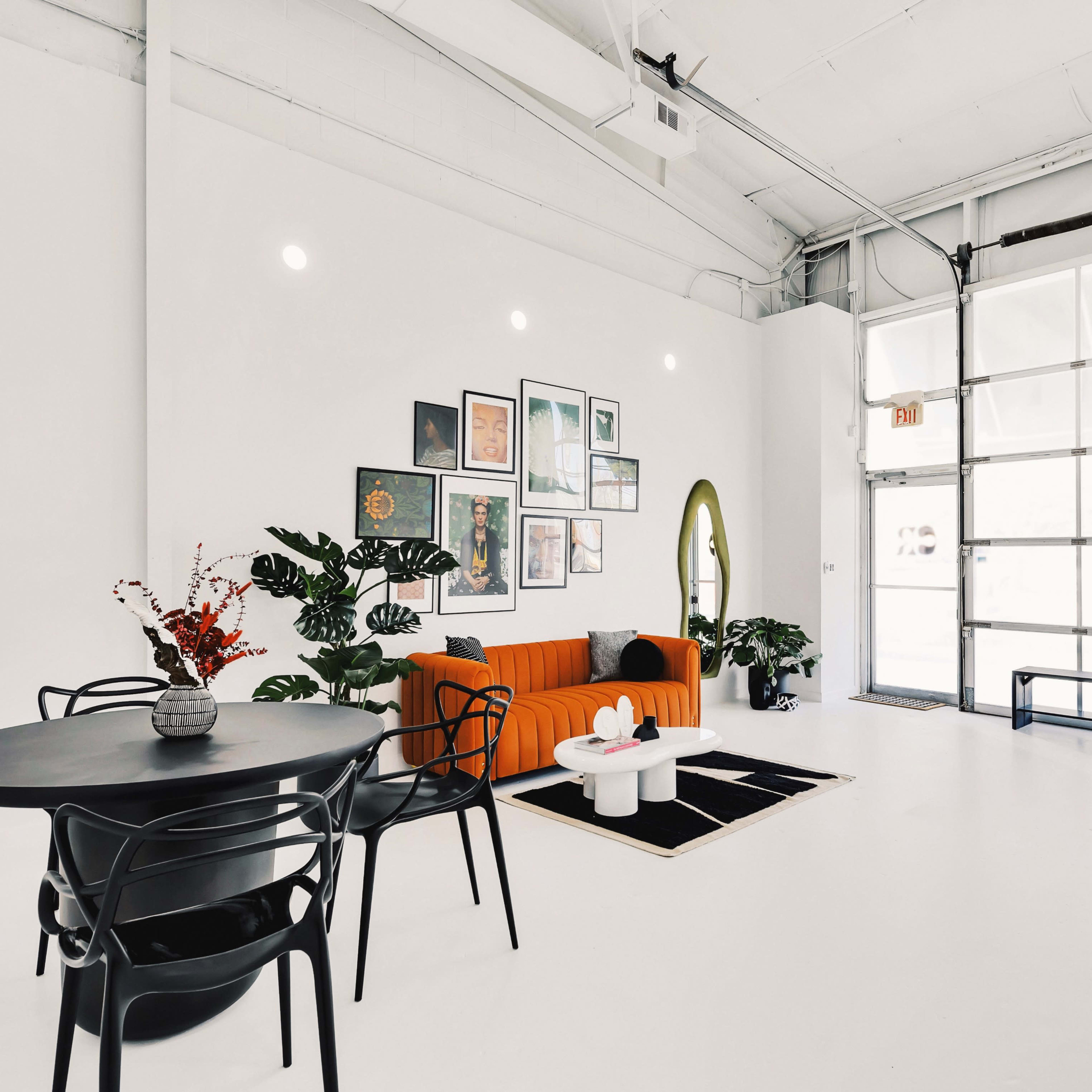 A bright, modern space featuring an orange couch against a white wall adorned with framed artwork, a black dining table with chairs, and a large garage door letting in natural light.