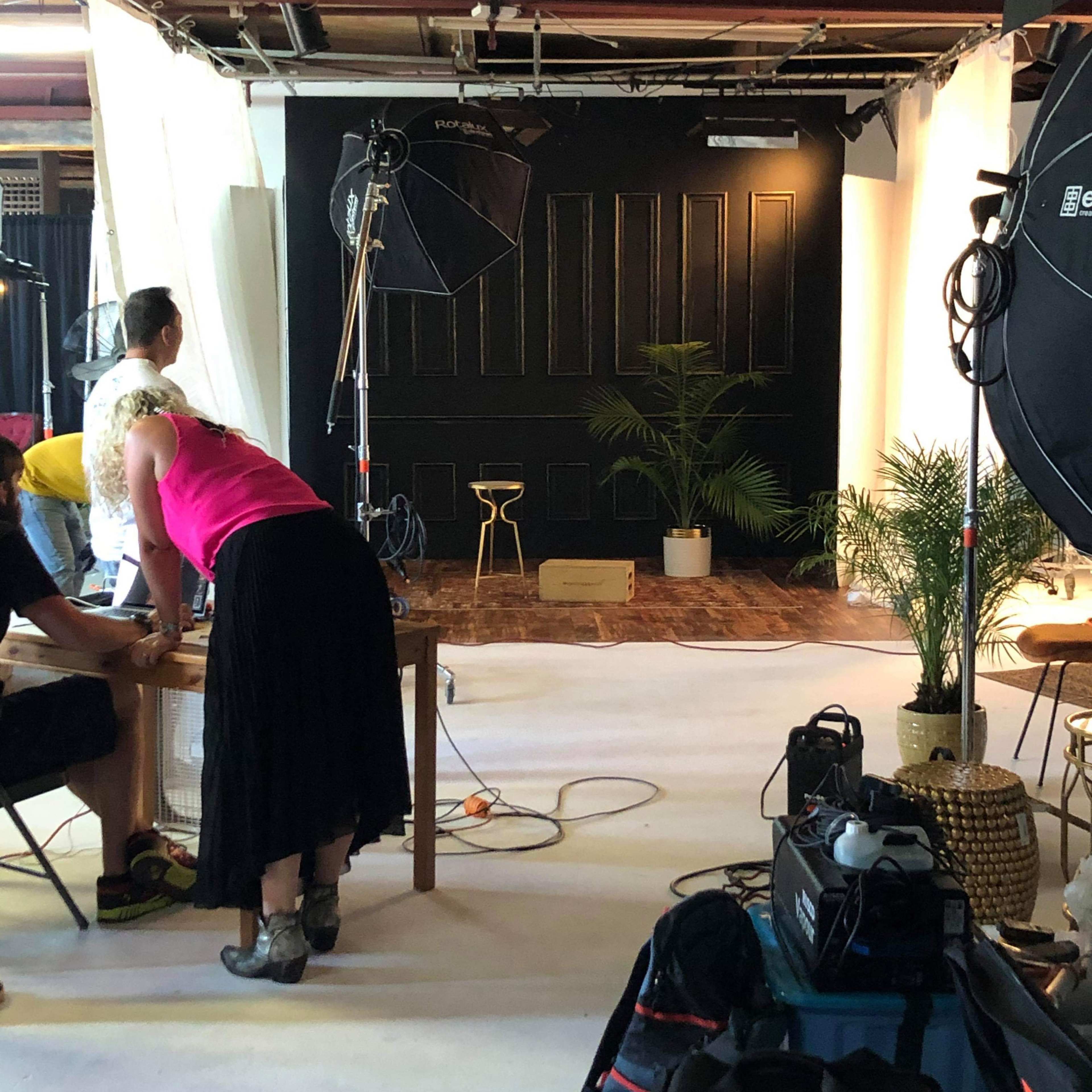 A photography studio setup features a black backdrop, lighting equipment, a stool, and several plants, with two people preparing for a shoot.