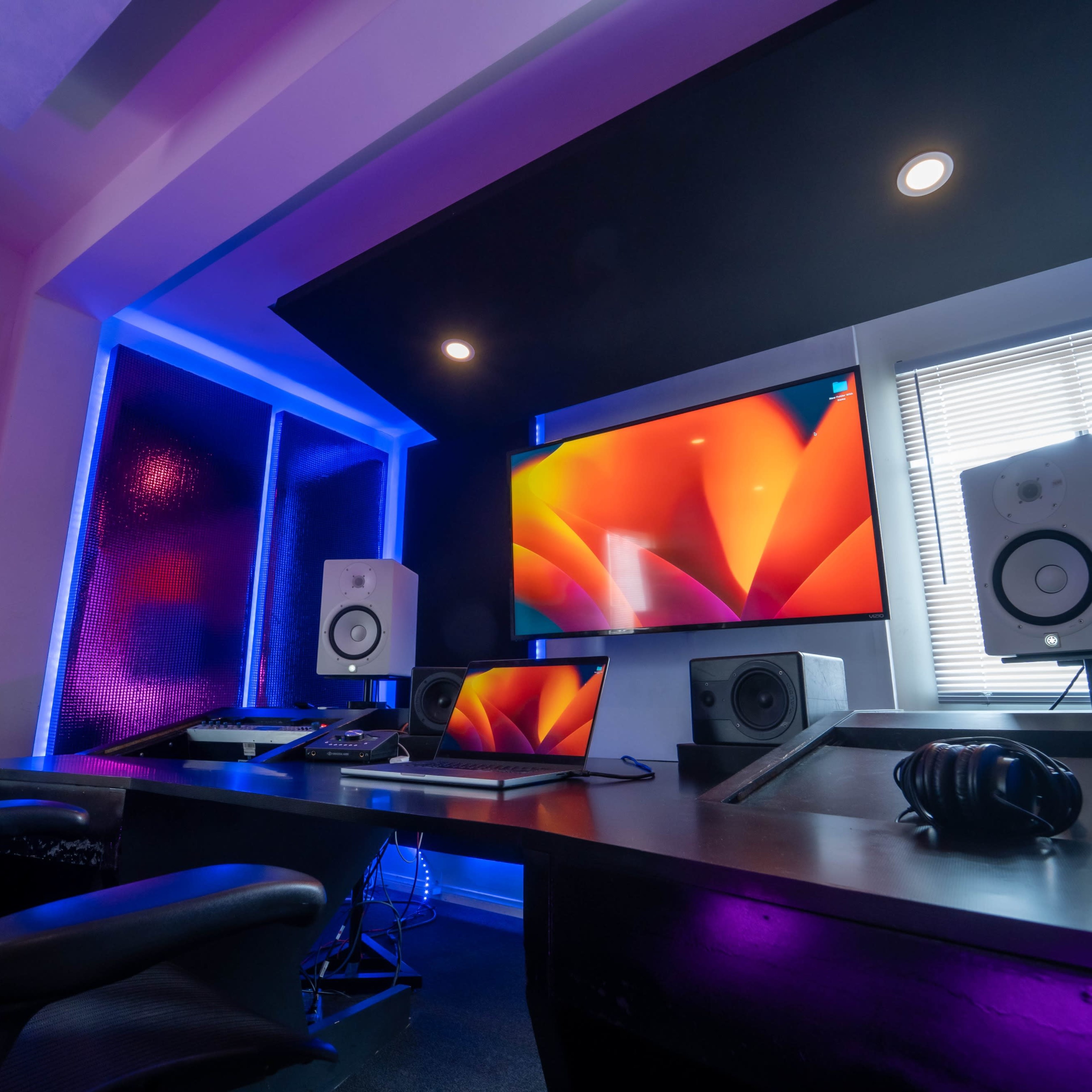 A modern music production studio featuring a large screen, studio monitors, and a sleek desk setup illuminated by colorful lighting.