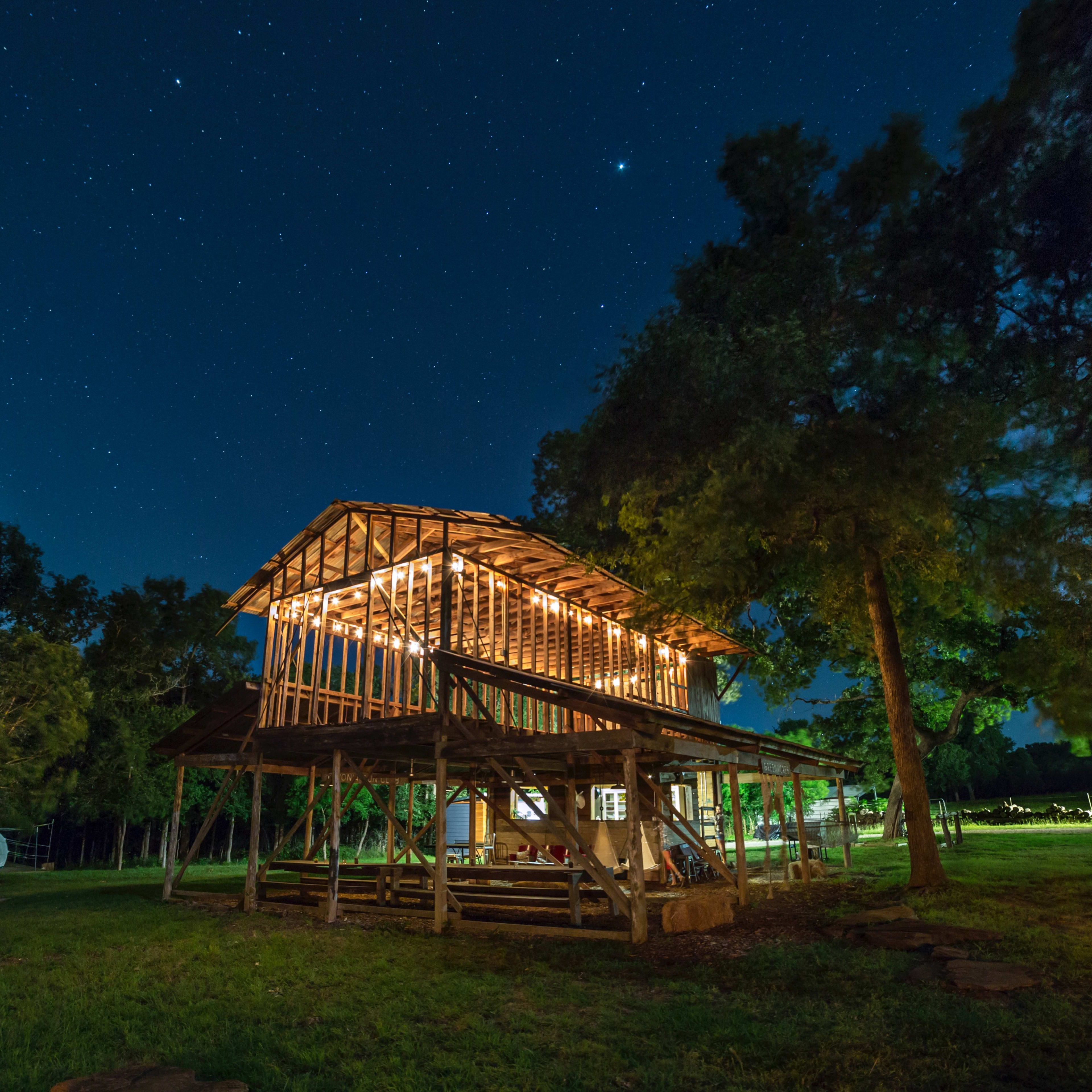 A partially constructed wooden structure is illuminated by string lights under a starry night sky, surrounded by trees.