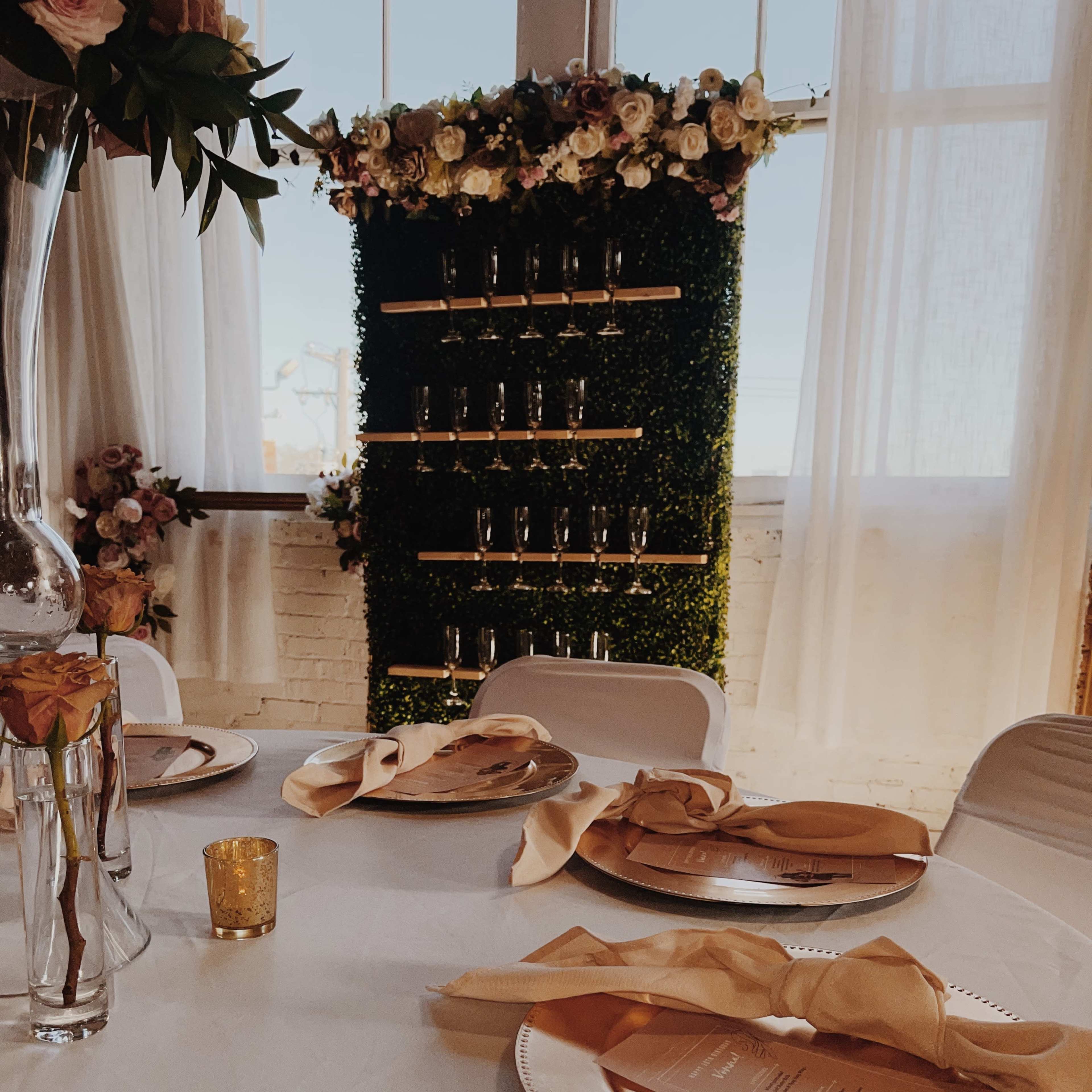 A elegantly set dining table with golden plates, napkins, and a backdrop of greenery adorned with flowers.