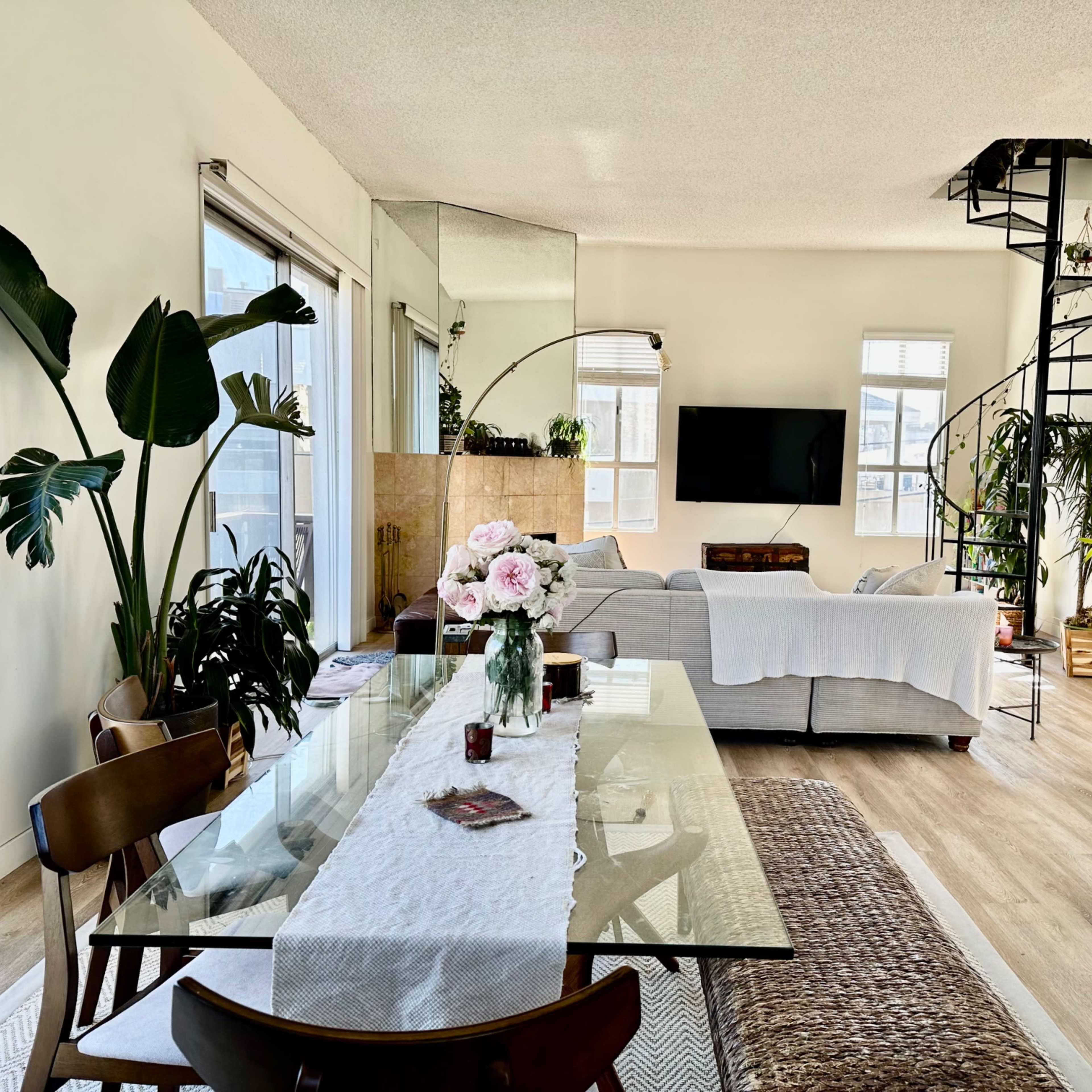 A bright, open living space features a dining area with a glass table and plants, a cozy seating area with a television, and a spiral staircase leading to an upper level.