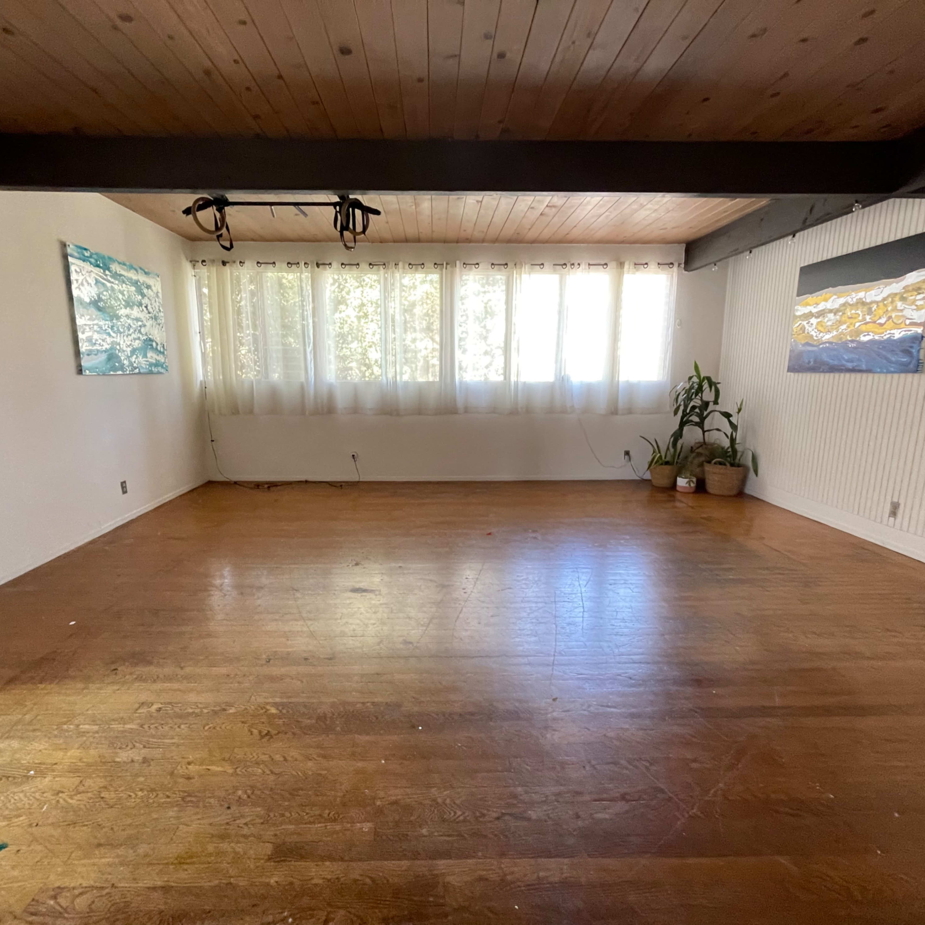 A spacious, empty room with wooden flooring, large windows covered by light curtains, and two artworks hanging on the walls.