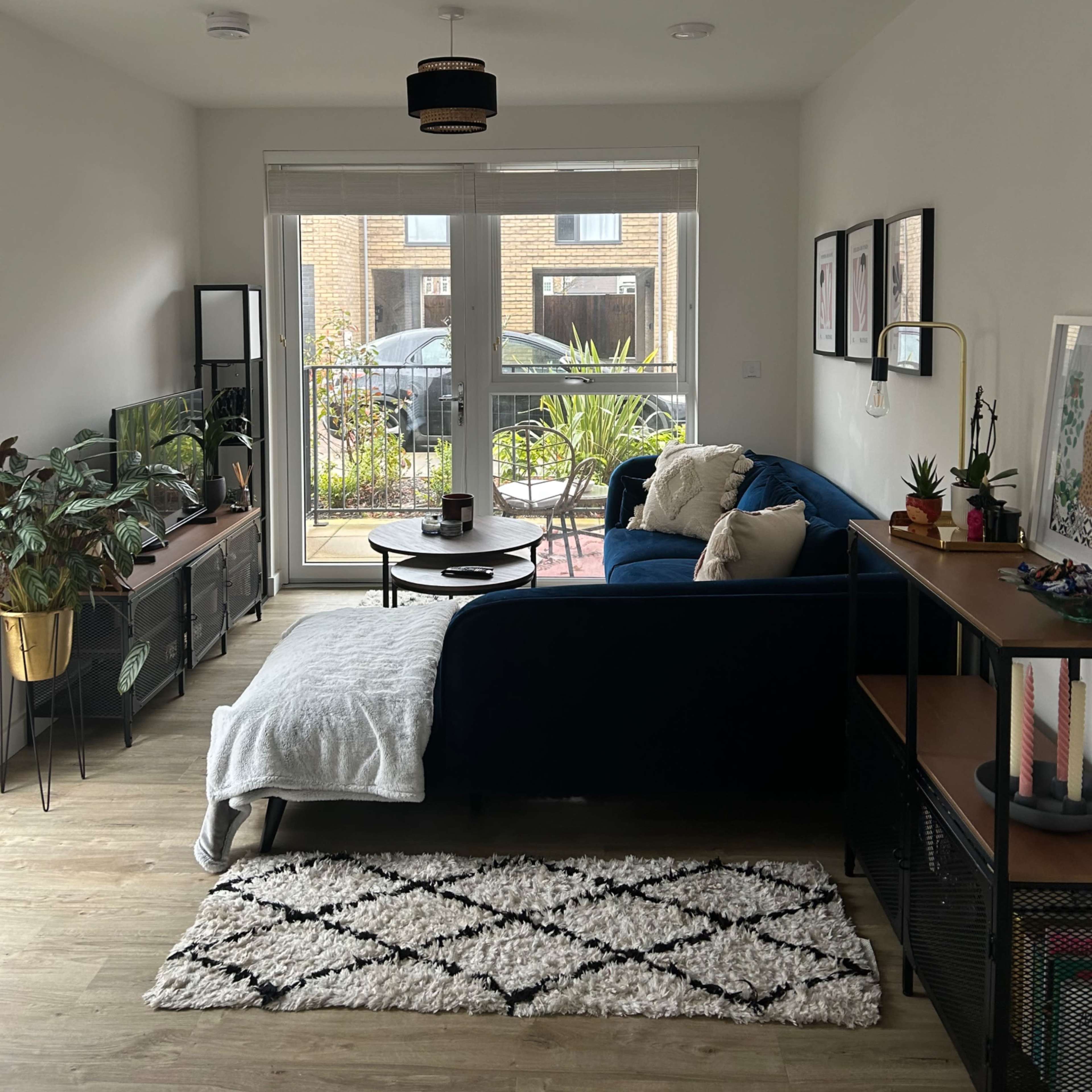 The living room features a blue sectional sofa, a round coffee table, a TV unit with plants, and large windows overlooking an outdoor area.