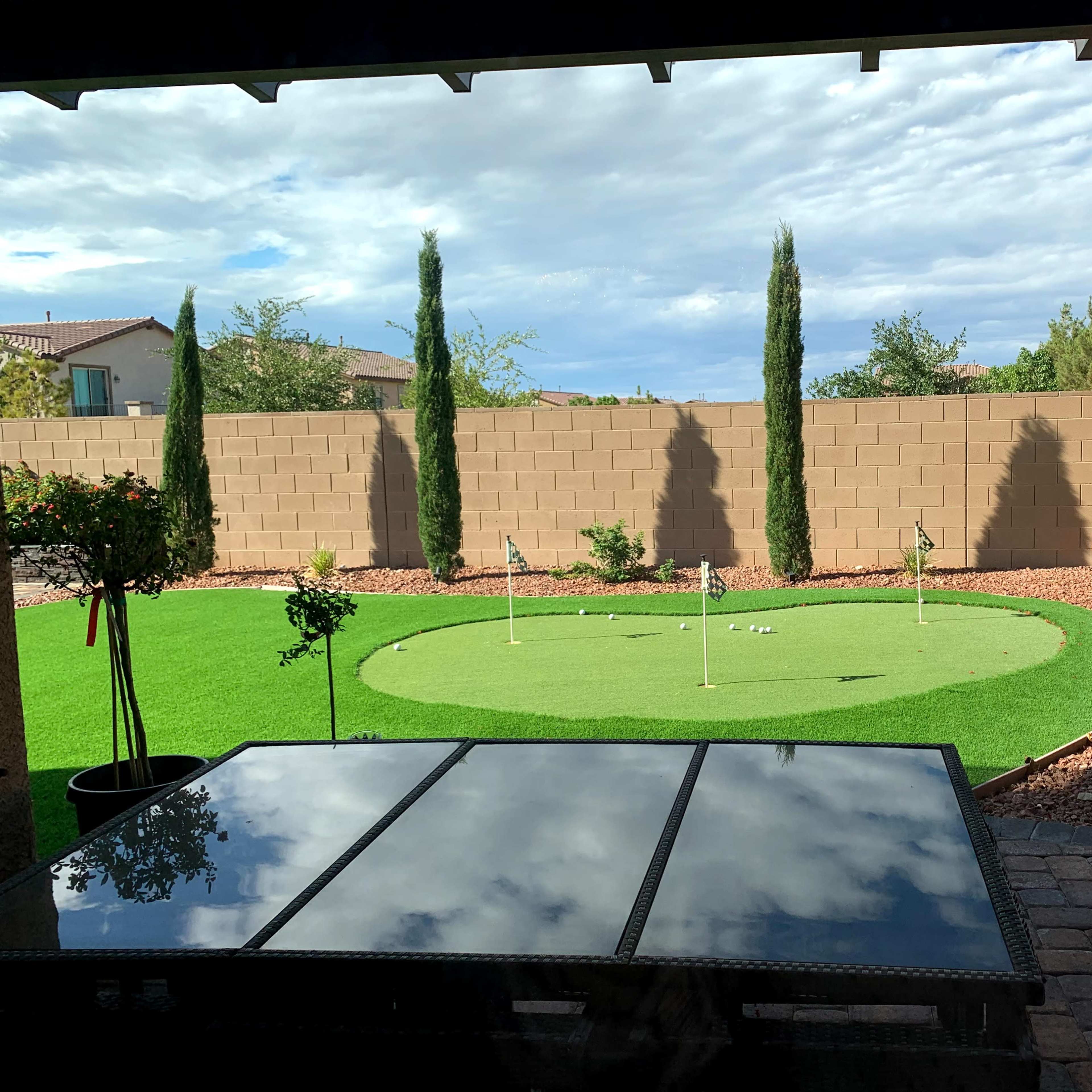 A backyard with a putting green surrounded by low shrubs, a stone path, and a covered patio.