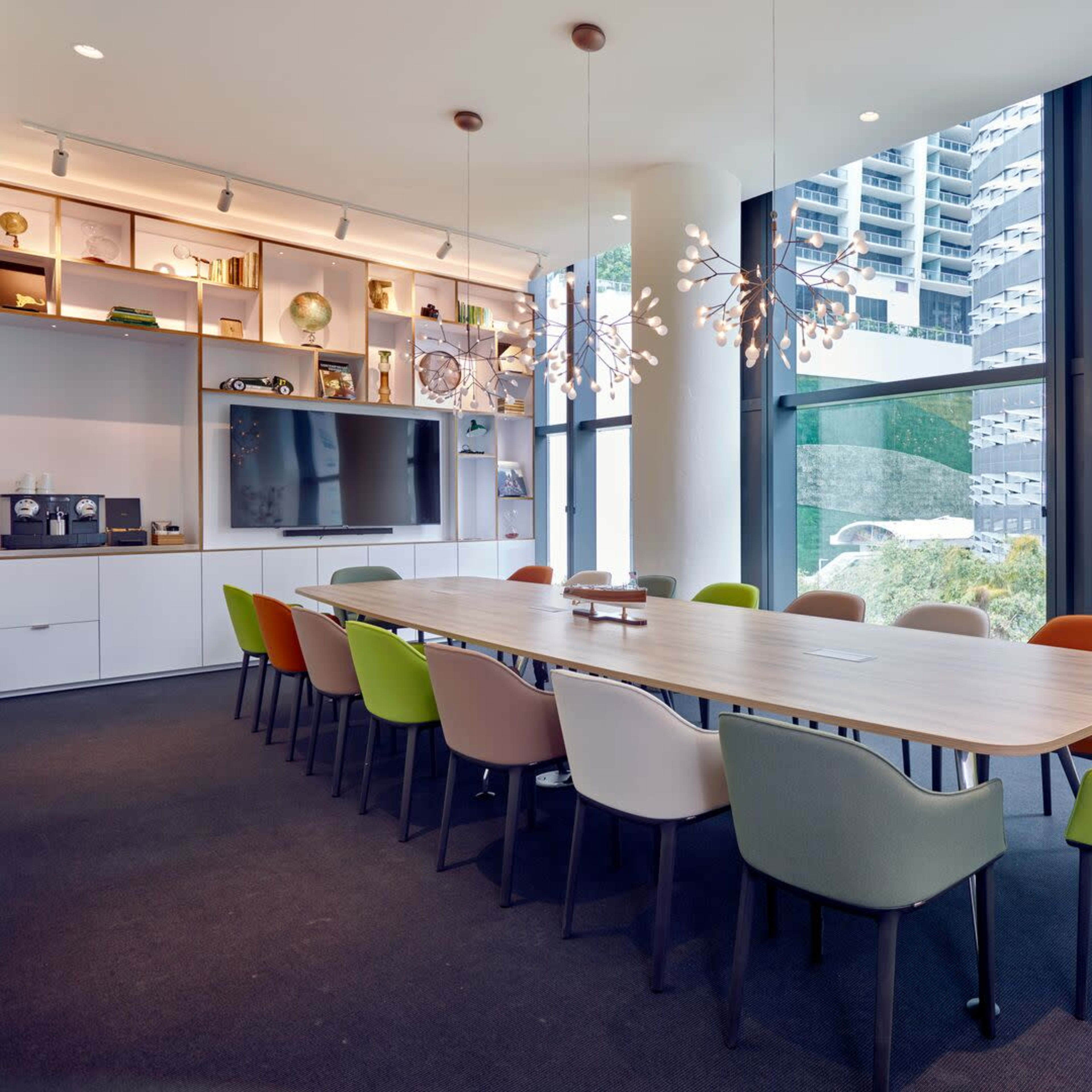 A long table with variously colored chairs is set up in a modern conference room with large windows overlooking a courtyard and building structure.
