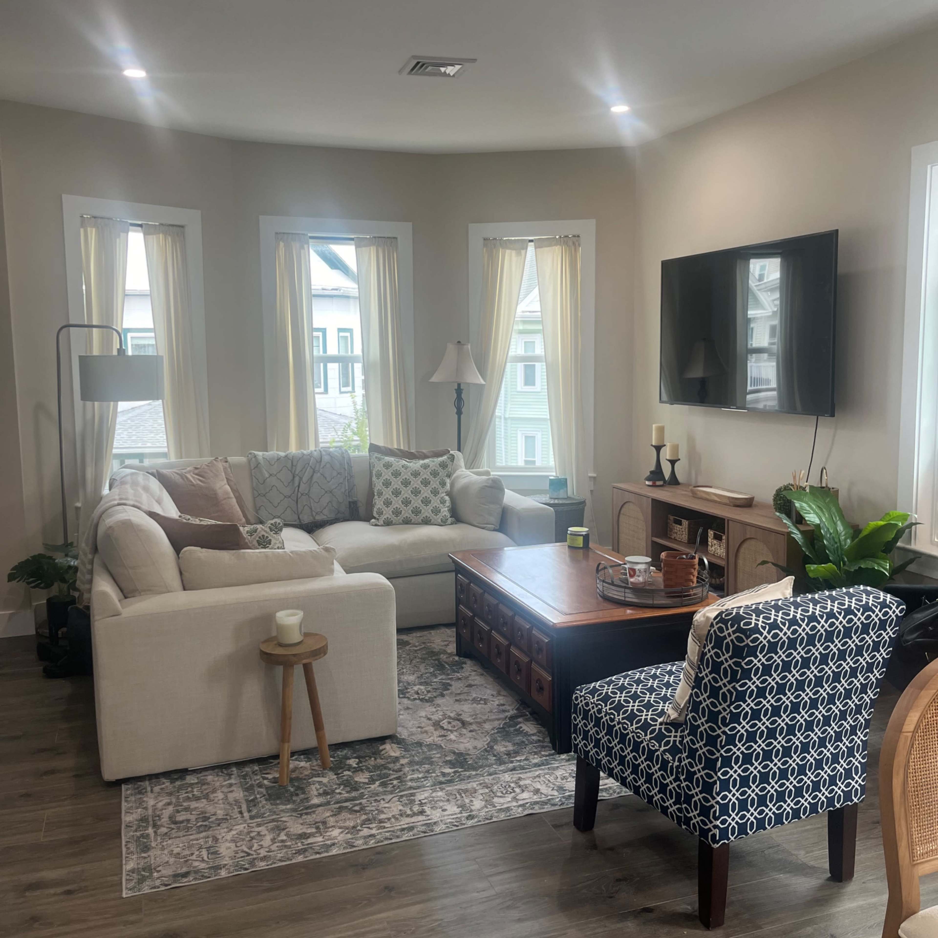 A bright living room featuring a white sofa, a dark wooden coffee table, a patterned chair, and large windows with light curtains.