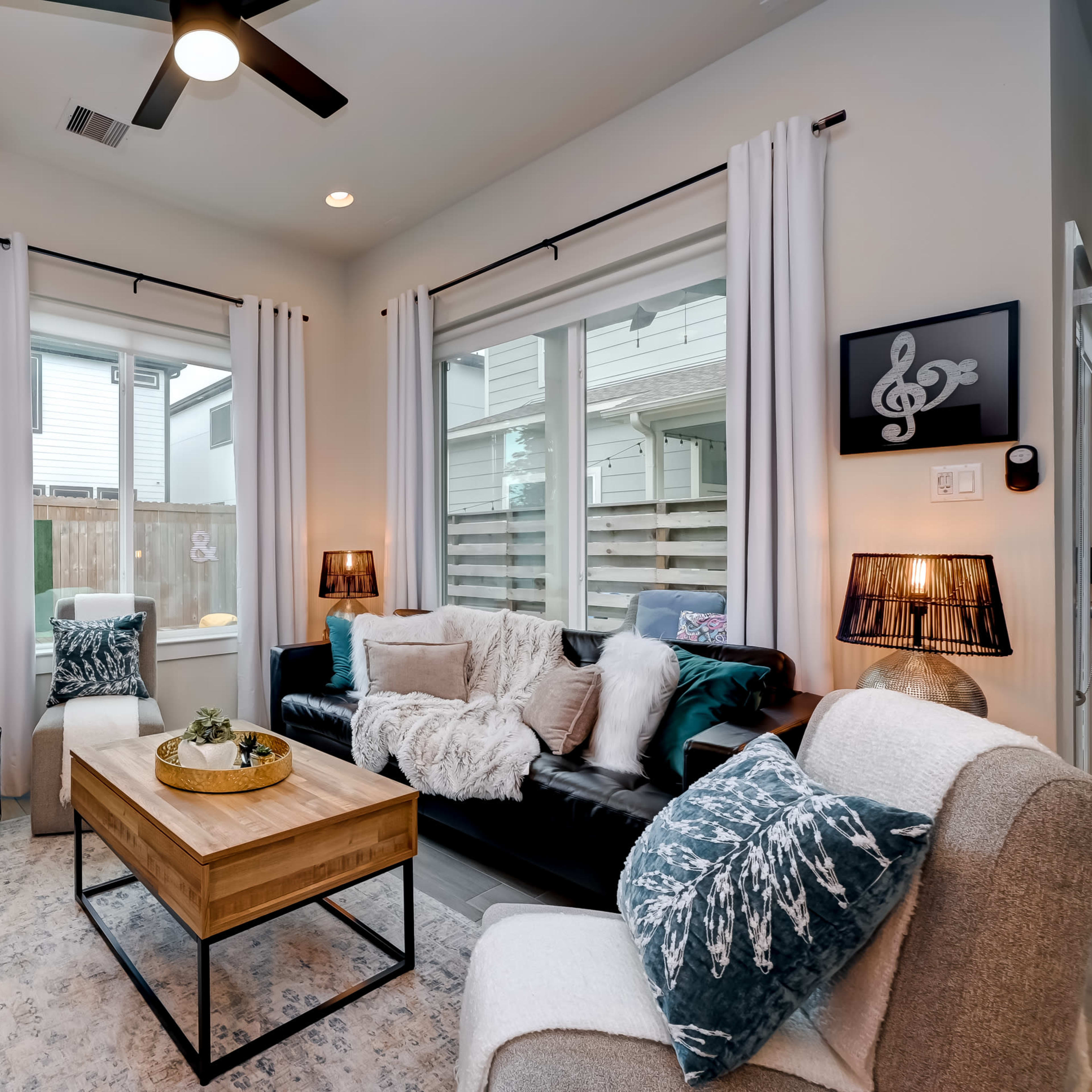 A modern living room features a black sofa, a wooden coffee table, and large windows with white curtains, decorated with plants and a textured area rug.