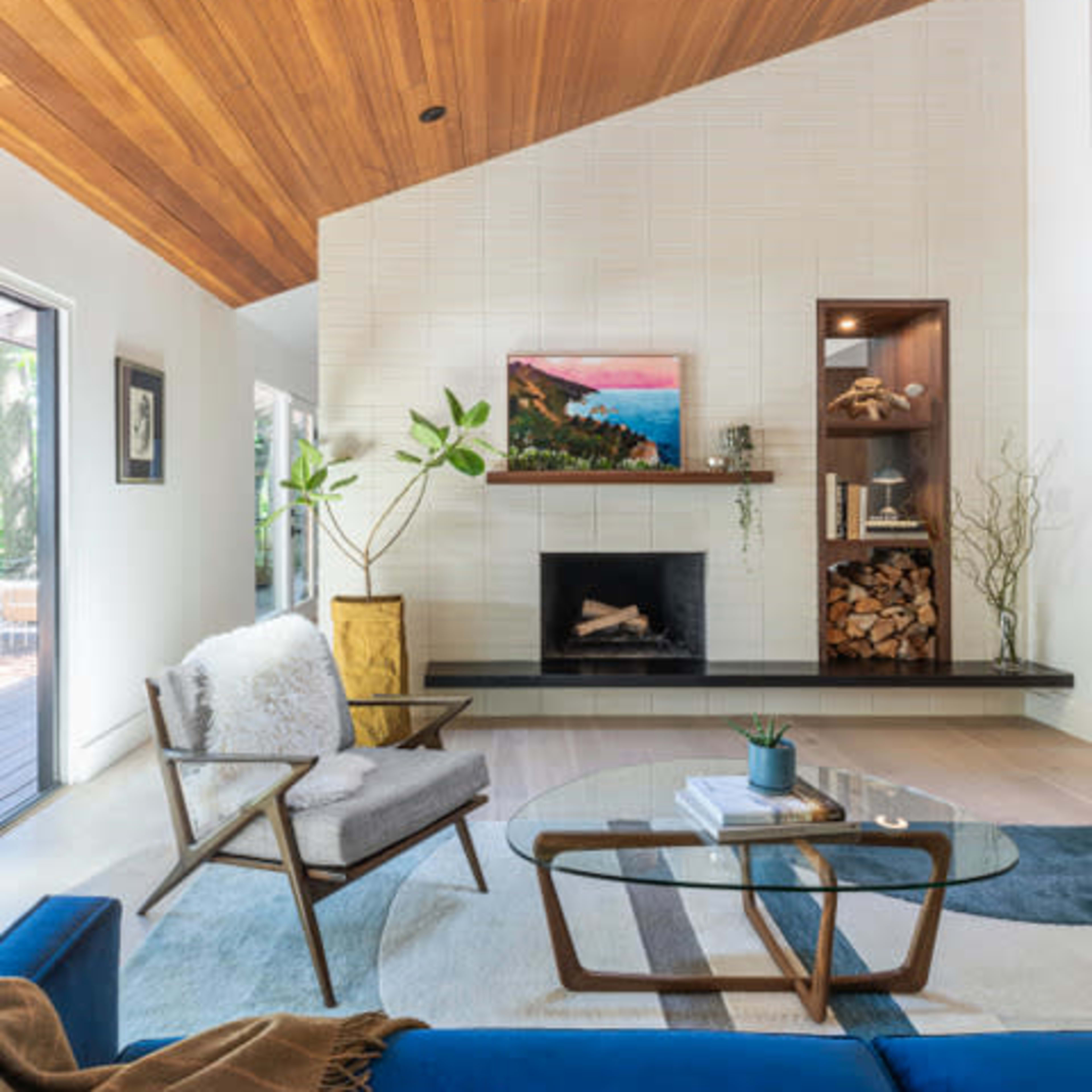 A modern living room featuring a wooden ceiling, a fireplace with a mounted painting above it, and a combination of plants and decorative elements.
