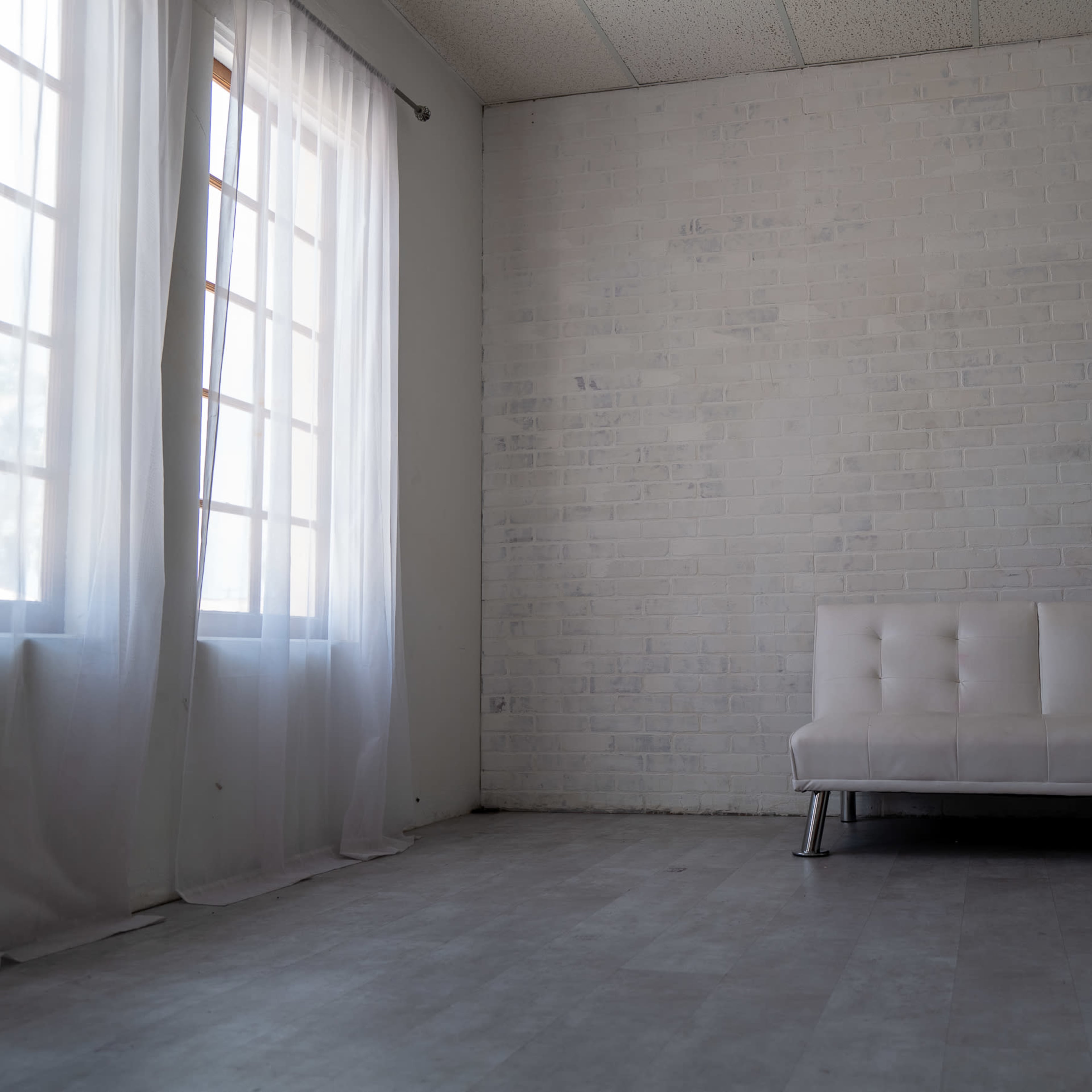 A minimalist room with a white brick wall, a white sofa, and large windows draped with sheer curtains.