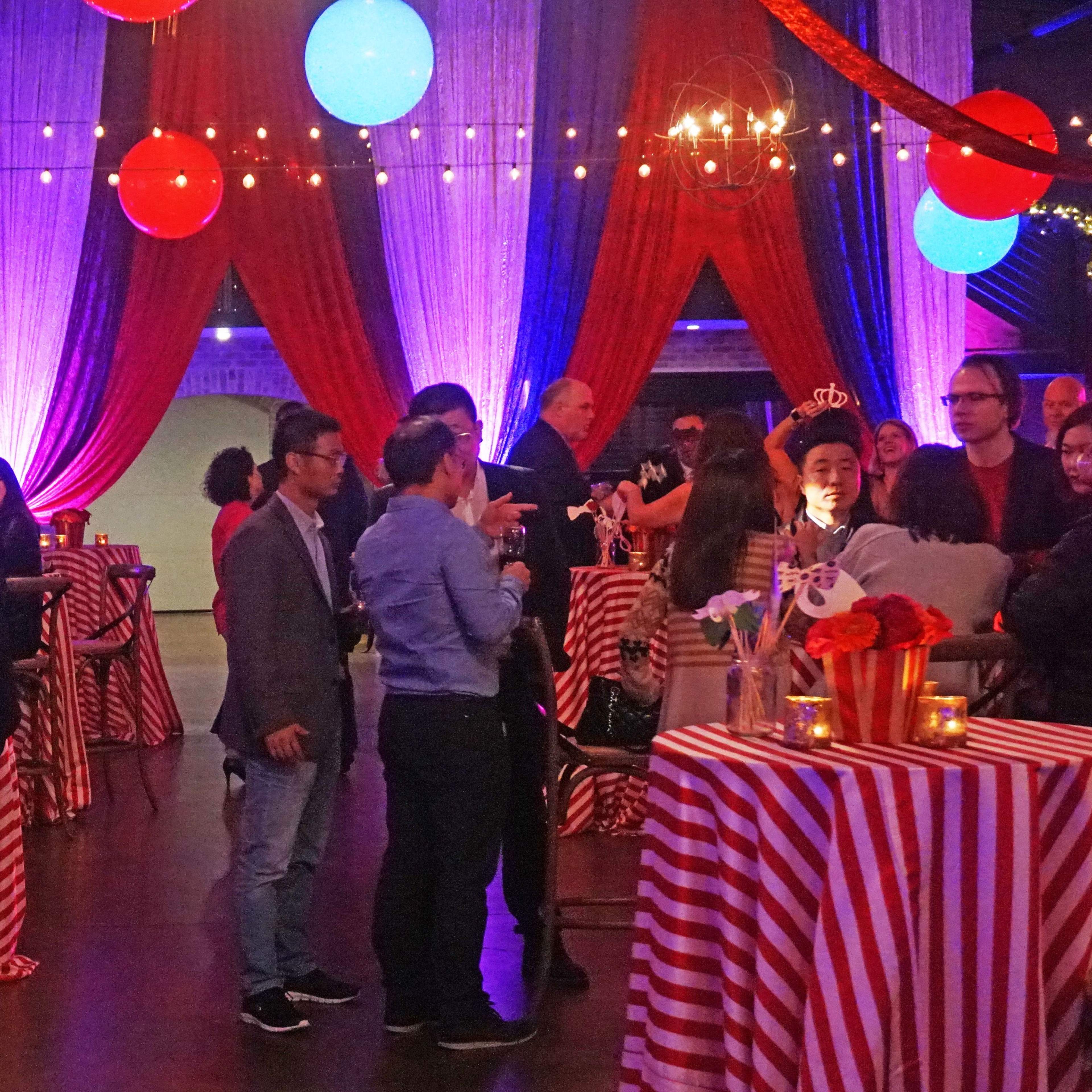 Guests socialize at tables with striped coverings in a festively decorated event space featuring colorful hanging lights and draped curtains.