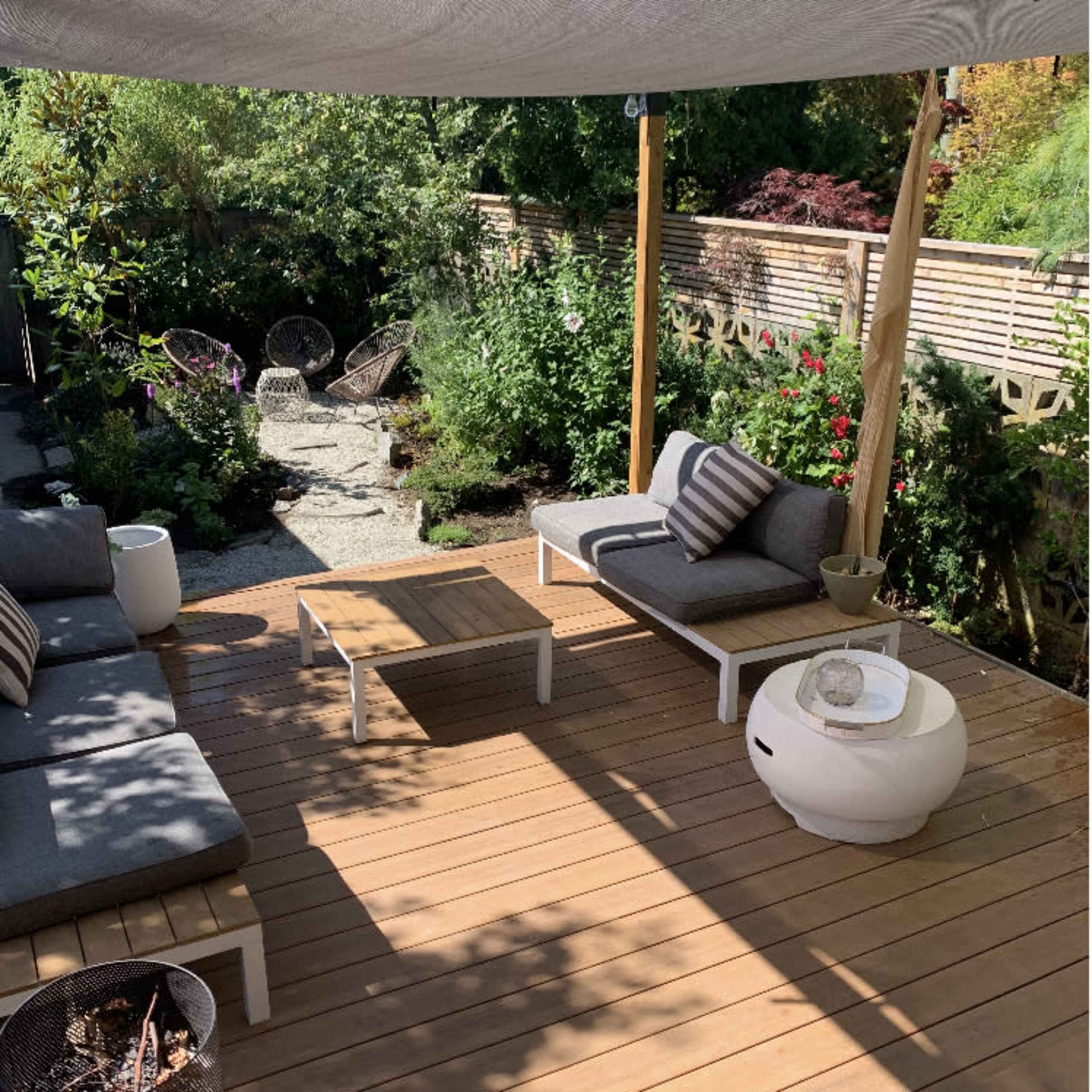 A wooden patio area with two lounge chairs, a coffee table, and a shaded seating arrangement surrounded by greenery and stone pathways.