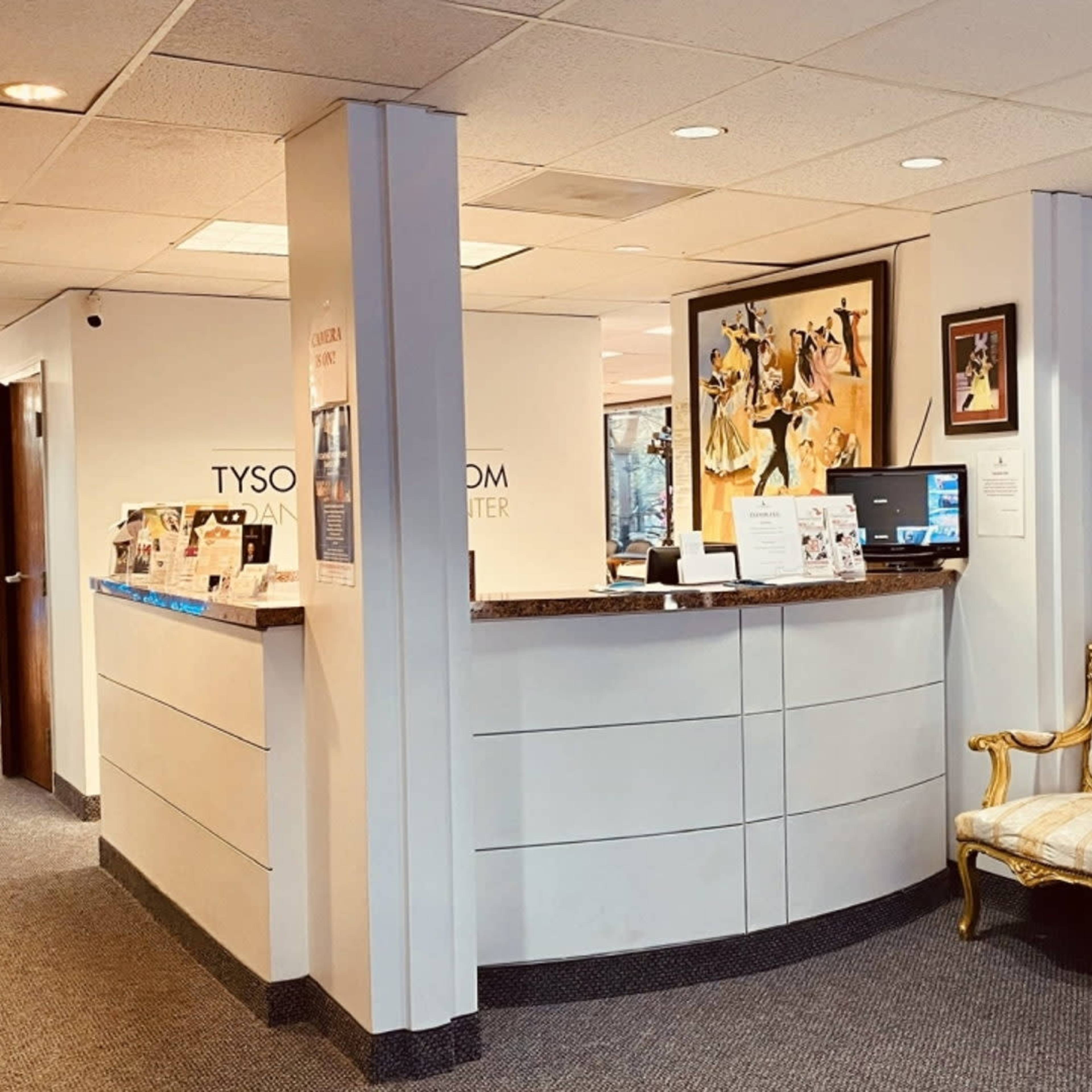 A reception area with a front desk, an ornate gold-accented couch, and a wall clock, all beneath bright overhead lighting.