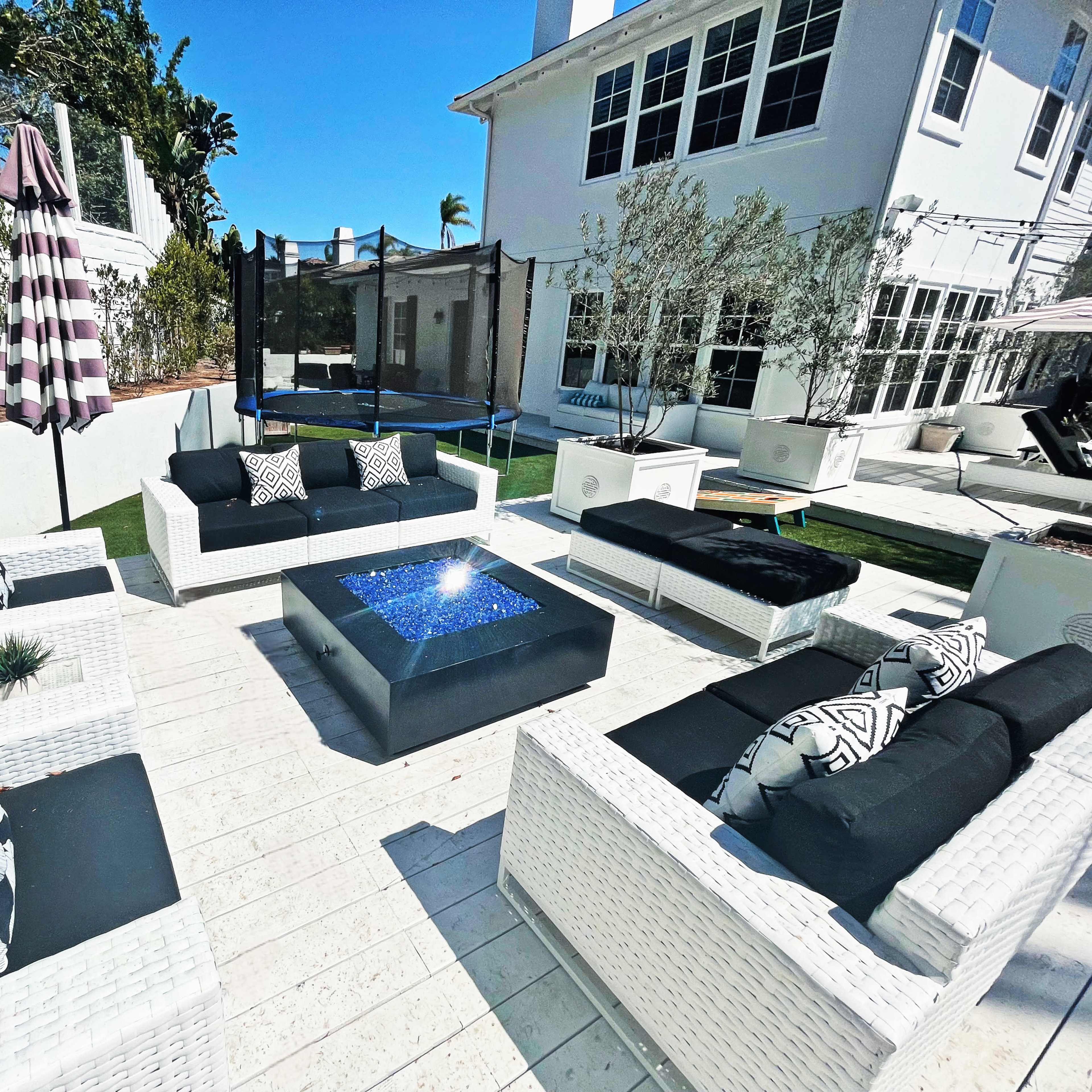 A patio area featuring white wicker seating arranged around a central fire pit, with a trampoline and a large house visible in the background.