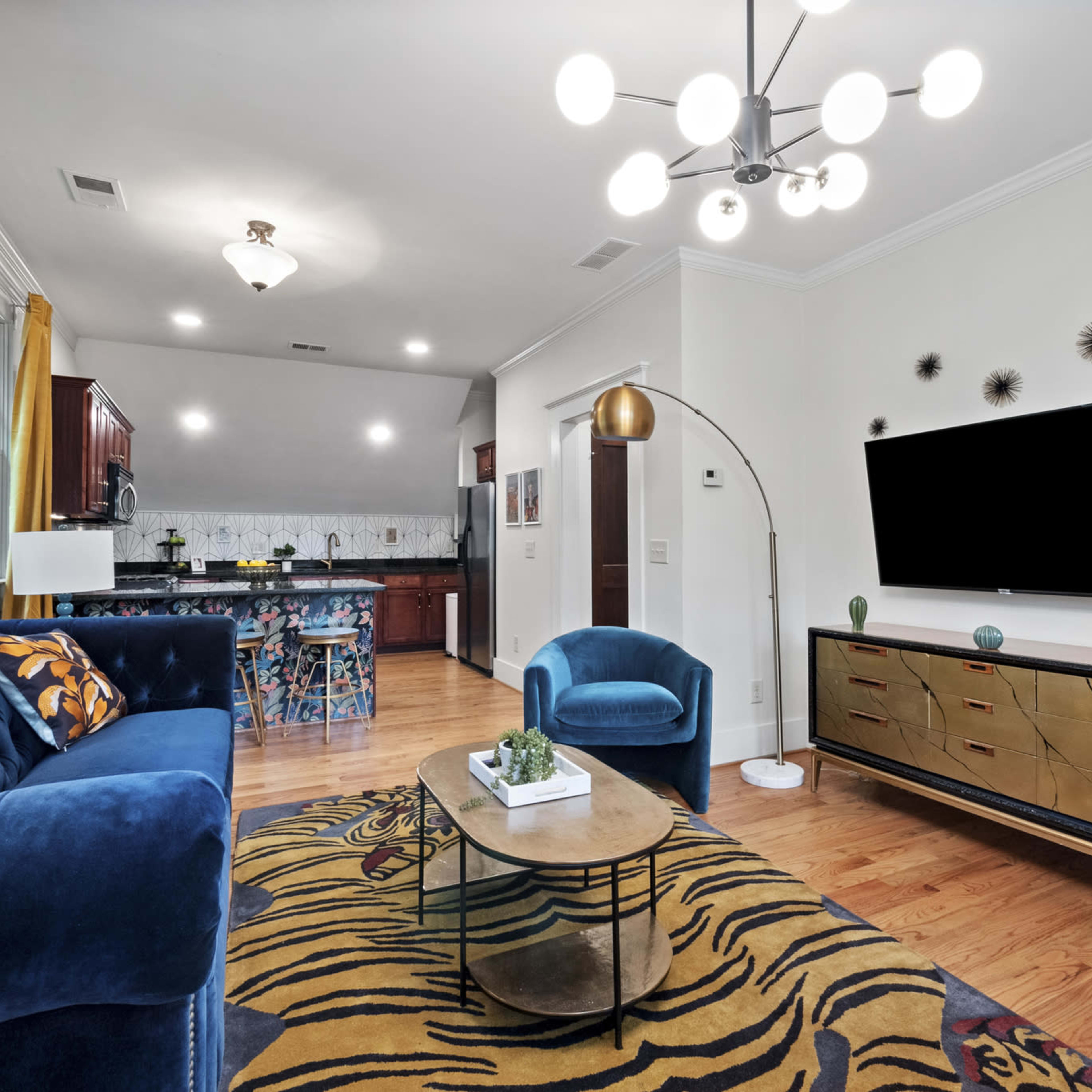 A modern living room featuring a blue velvet sofa, a gold-accented console table, and a large flat-screen TV mounted on the wall, complemented by a decorative yellow and black rug.