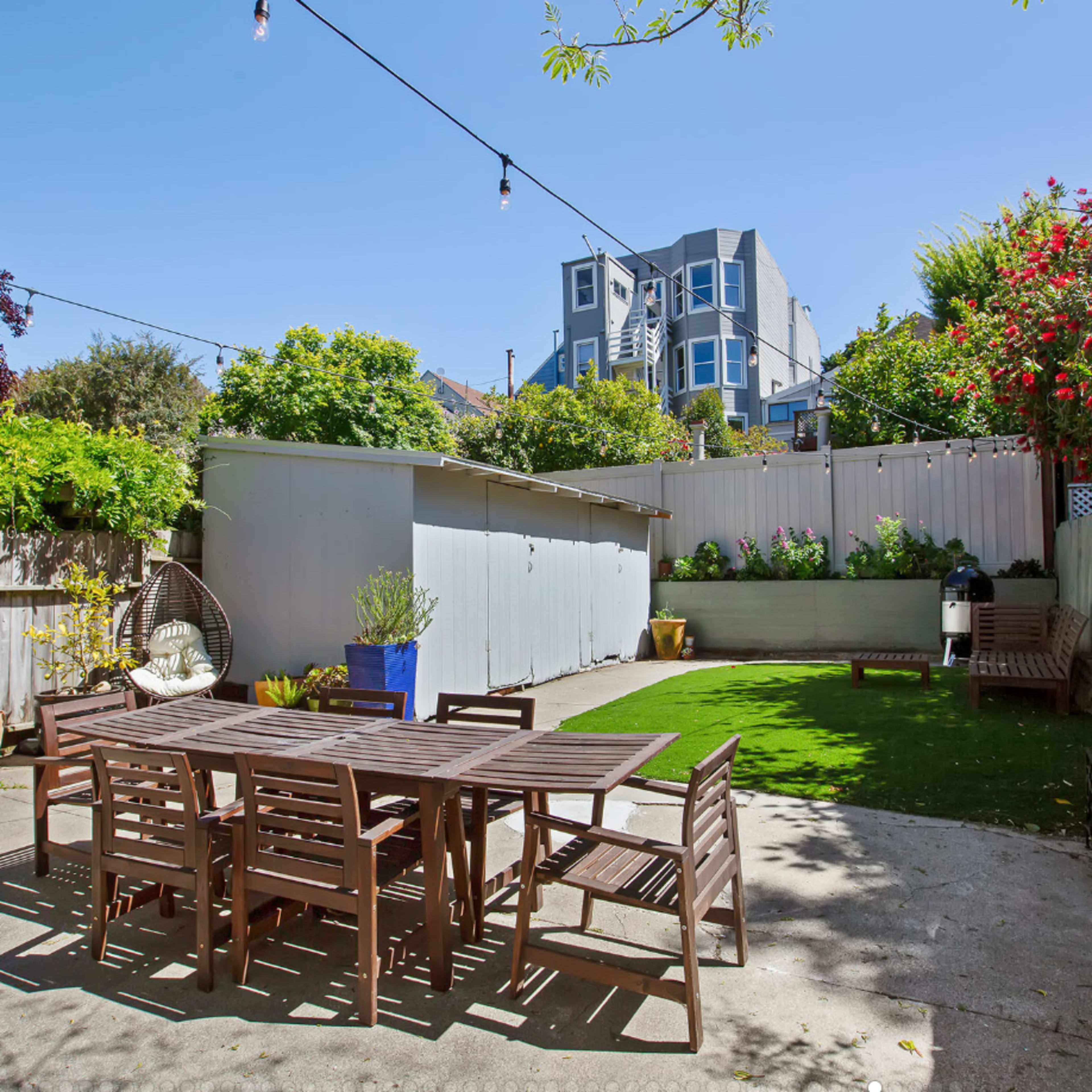 A backyard with a wooden dining table and chairs, surrounded by greenery and a garden shed in the background.