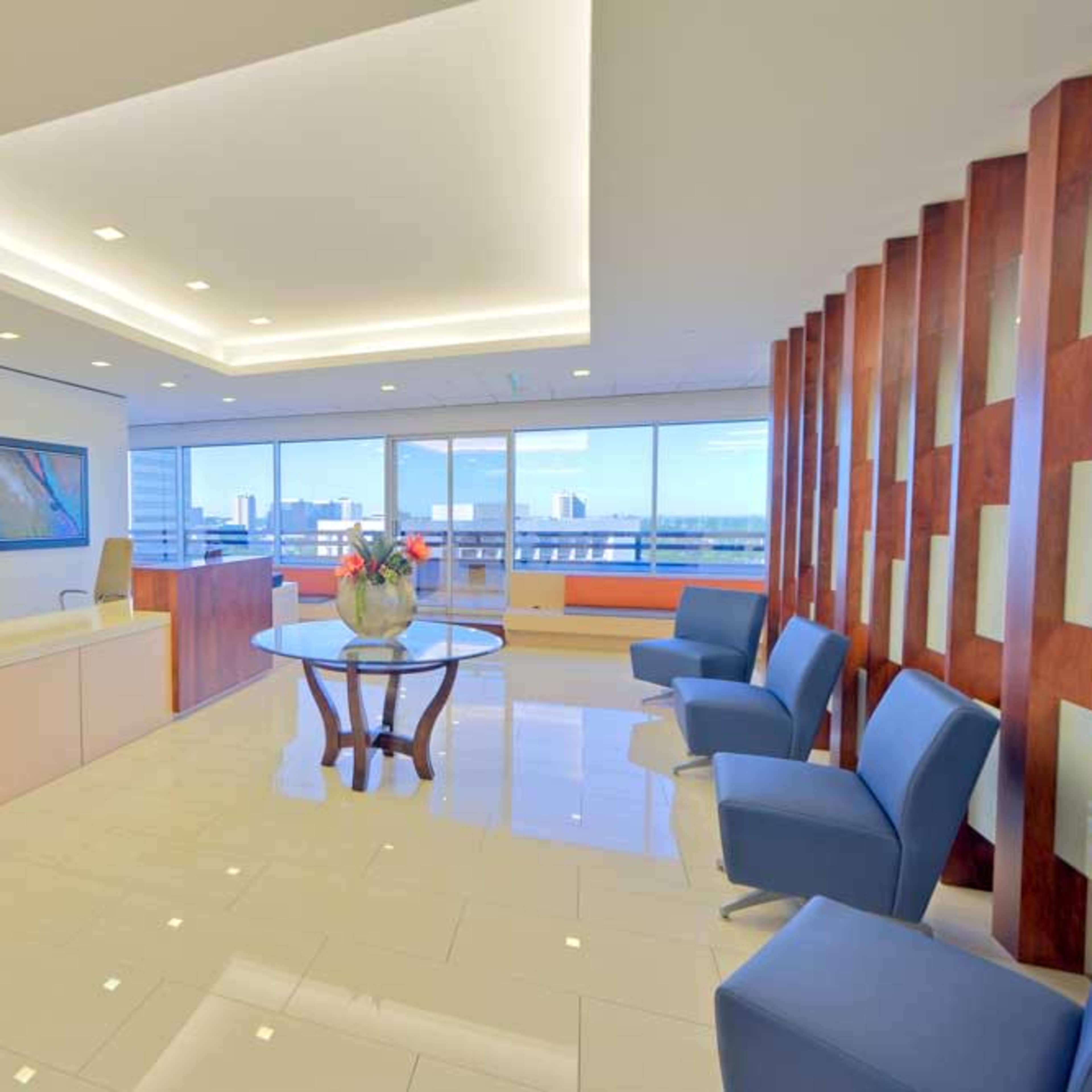 A modern office reception area with a front desk, two blue chairs, and large windows displaying a city skyline.