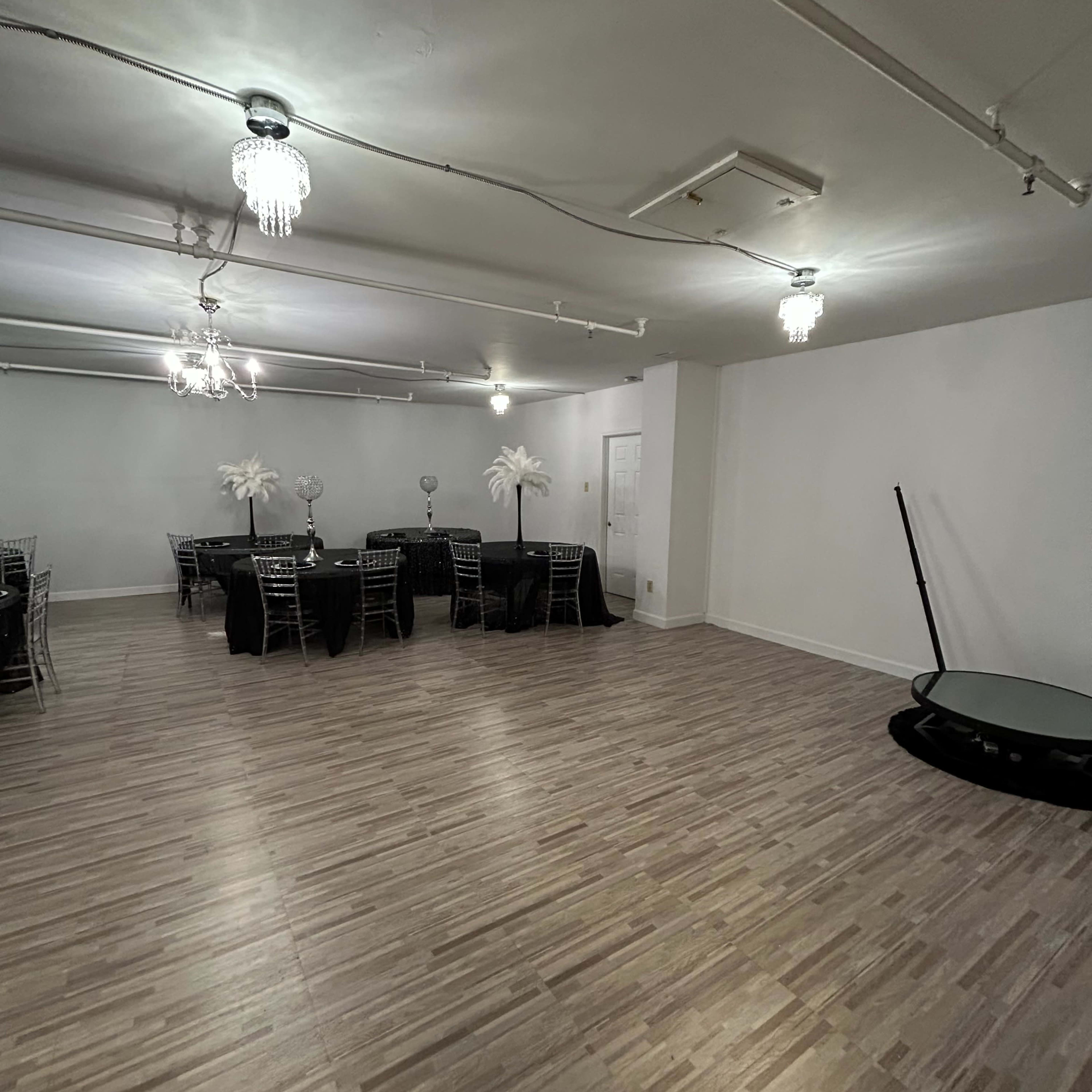 A spacious, empty event room with several black tables draped in white tablecloths and decorative centerpieces.