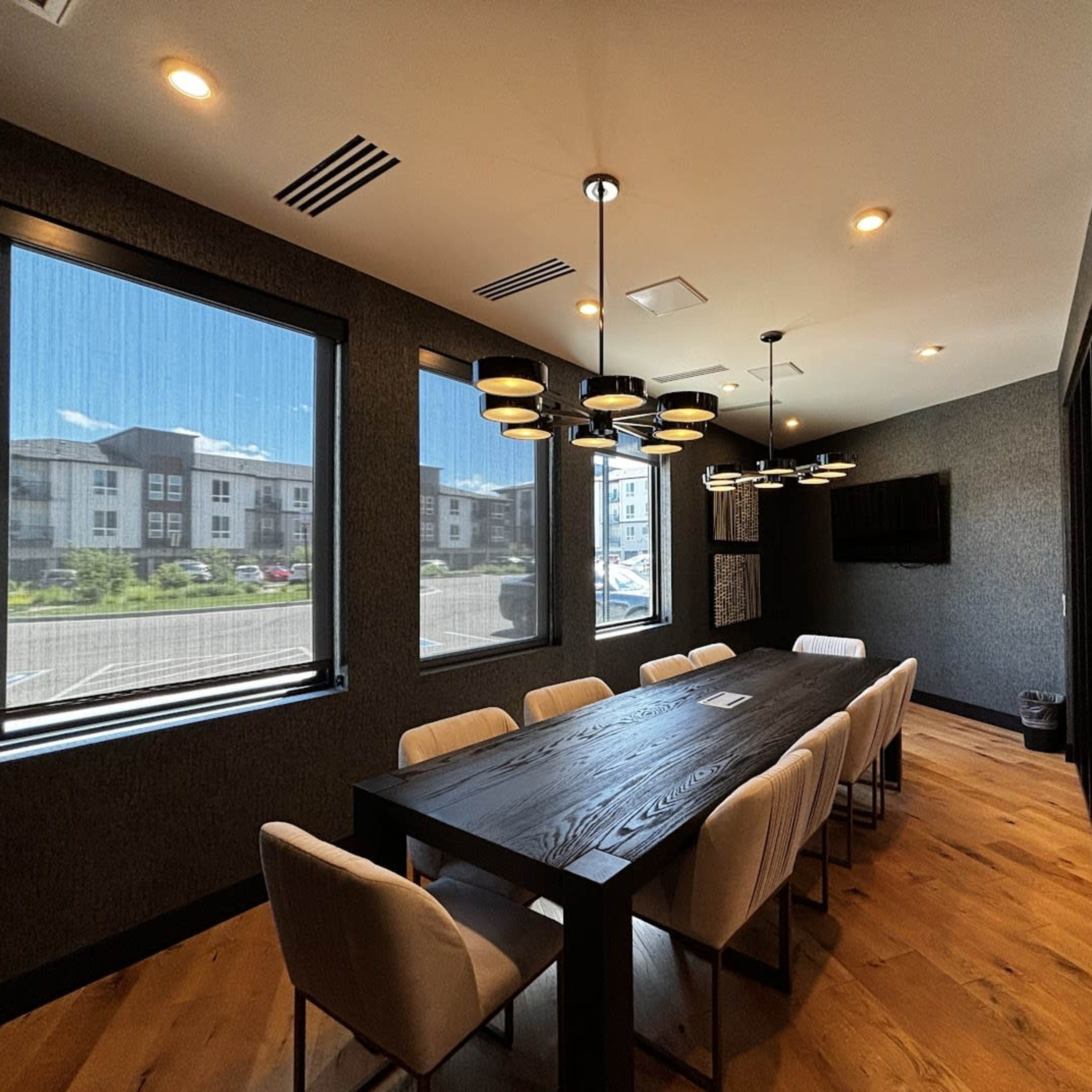 A modern conference room features a large black table surrounded by light-colored chairs, illuminated by pendant lighting and natural light through large windows.