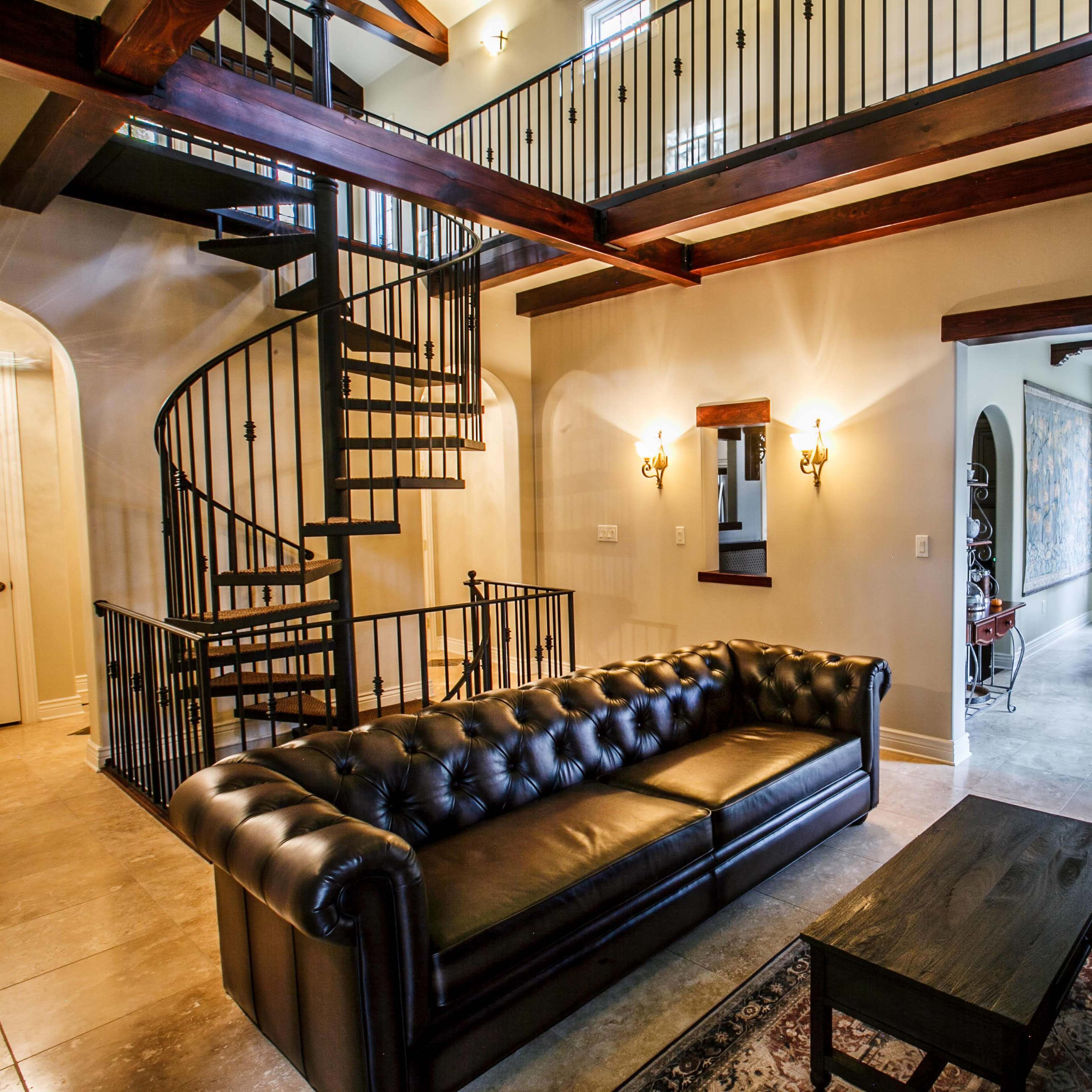 A spacious living area with a black leather sofa, a coffee table, and a spiral staircase leading to a second level, with a dining area visible in the background.