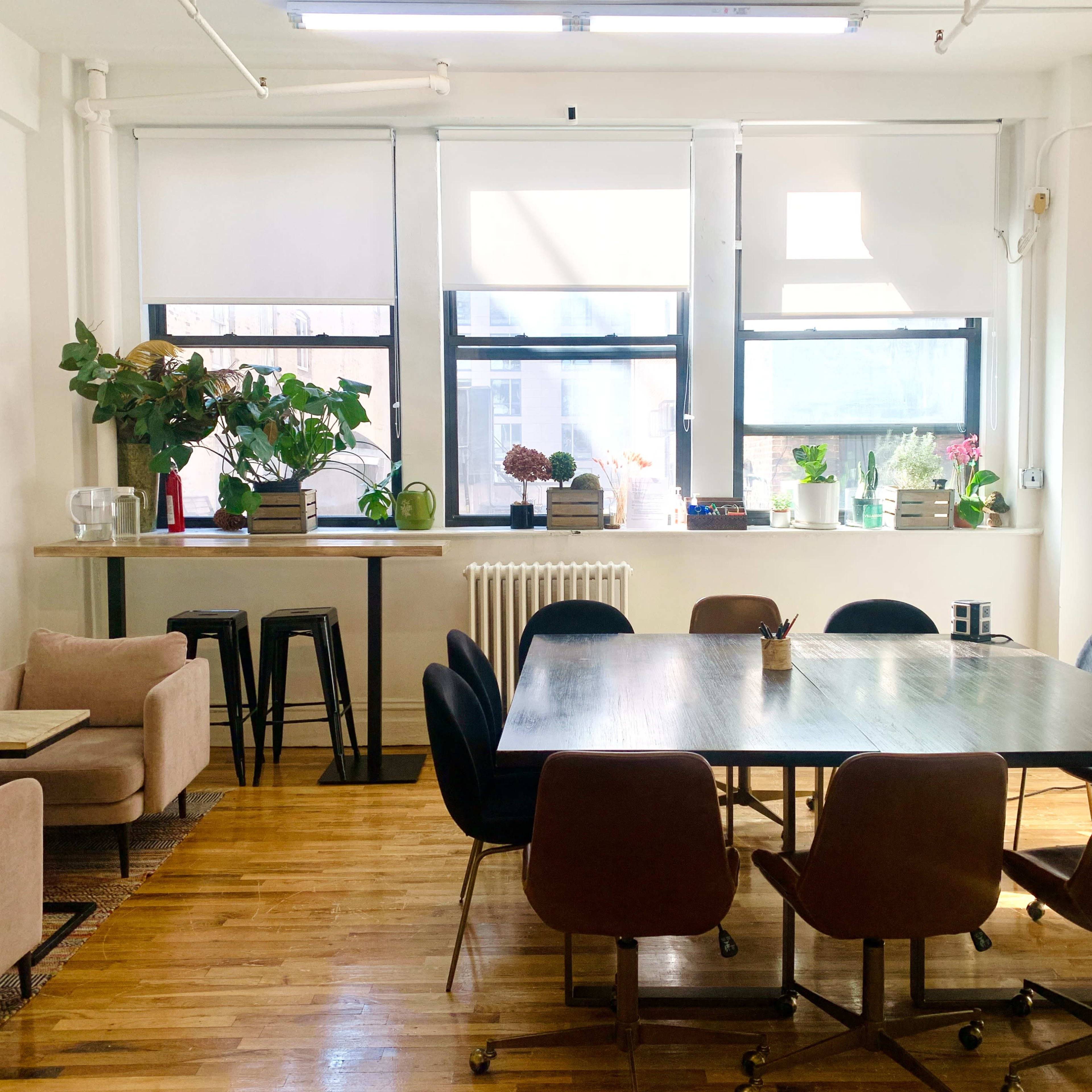 A bright, open room features a large table surrounded by chairs, with a small seating area and plants near the windows.