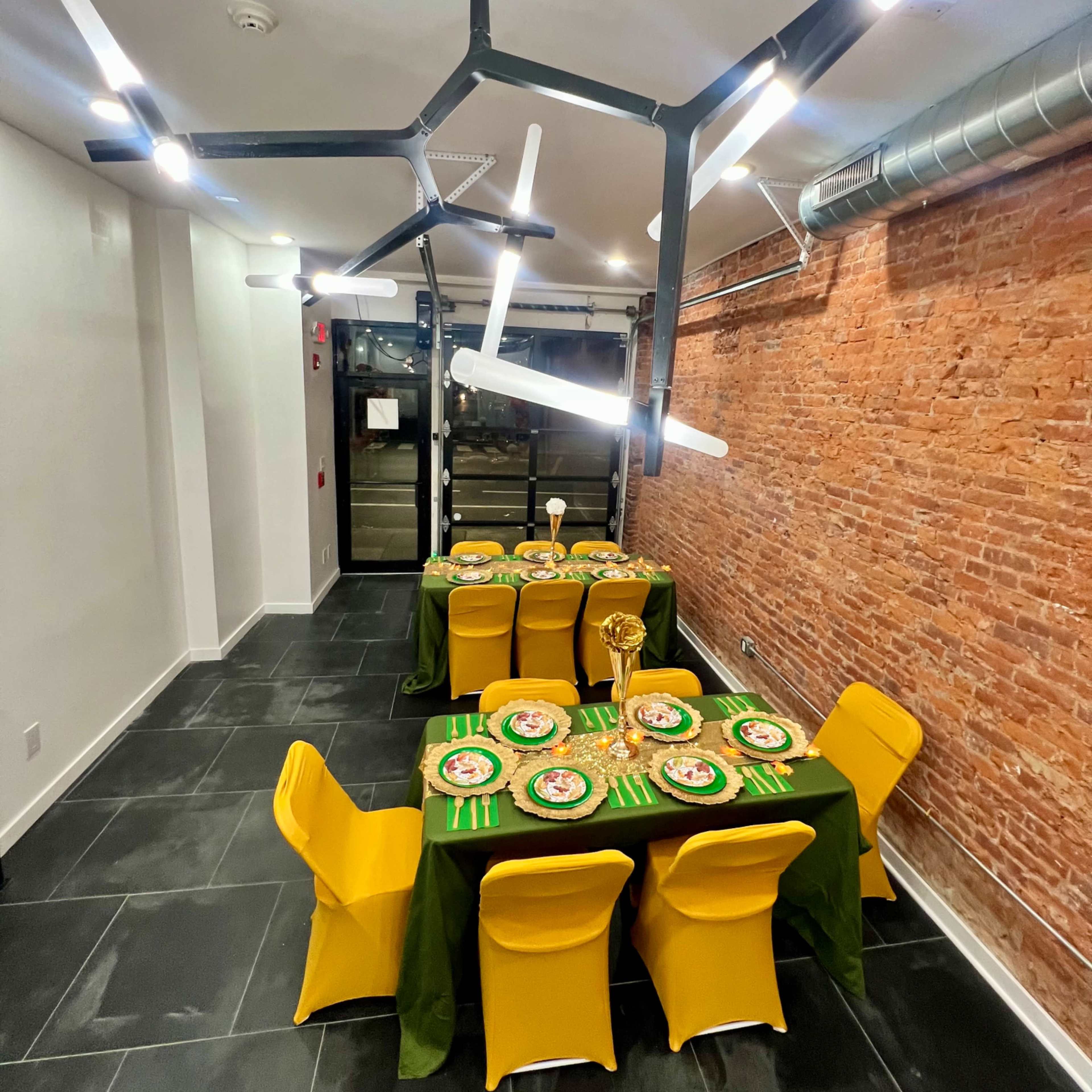 A dining area with two tables set for a meal, featuring green tablecloths and yellow chairs, illuminated by modern light fixtures against a backdrop of exposed brick walls.